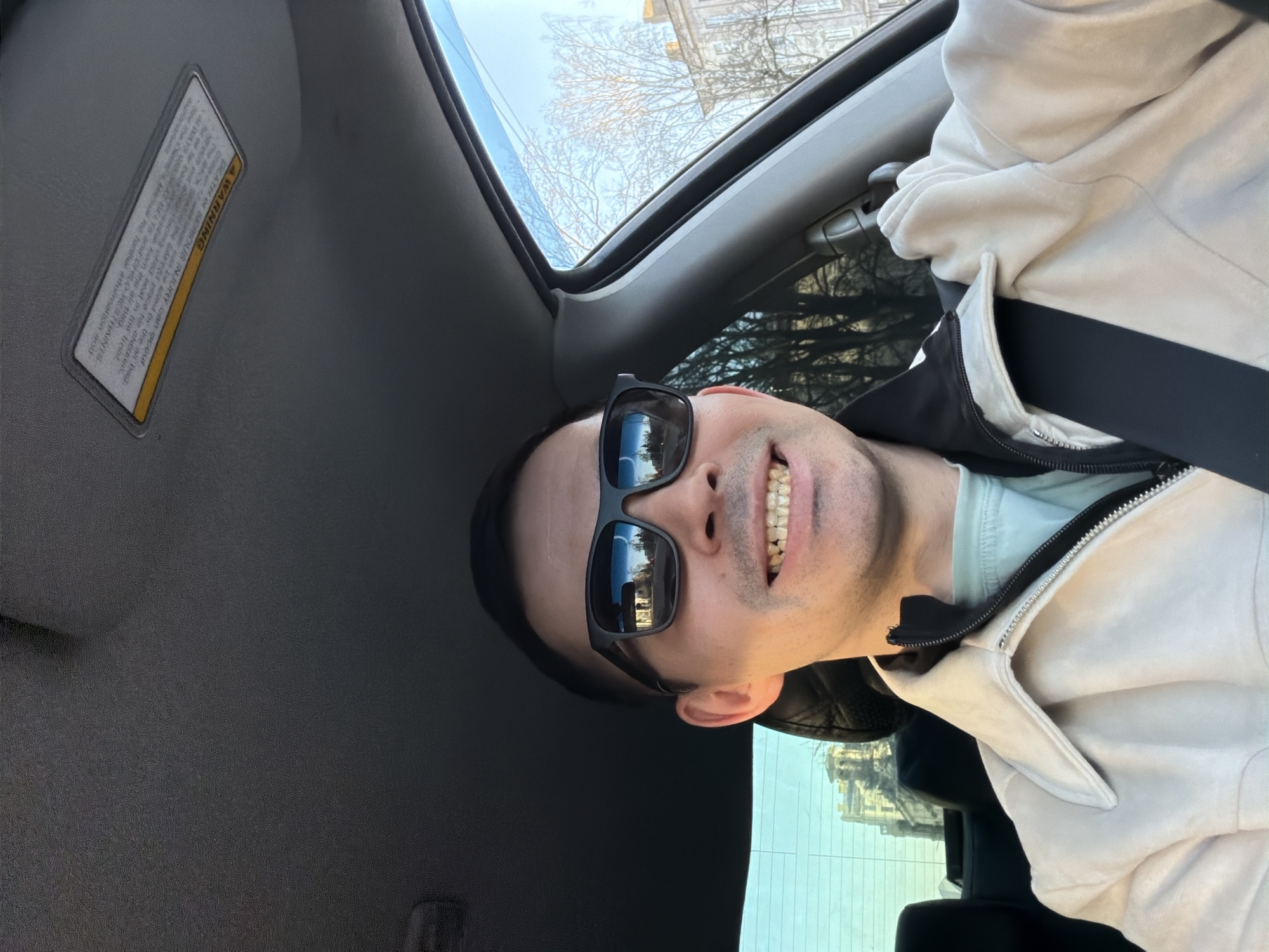 The image is a selfie taken from inside a car, showing a man smiling broadly at the camera. The man's head is tilted sideways, wearing black sunglasses that reflect a bright outdoor scene with buildings and bare trees. His mouth is open in a wide smile, revealing his teeth. He is dressed in a light-colored, possibly off-white or beige, zipped jacket worn over a darker layer, with a light blue or mint-colored shirt visible beneath.
The setting is the interior of a vehicle, likely a car, with the man appearing to be in the front passenger or driver's seat. Above him, a car sunroof reveals a clear blue sky and leafless trees, suggesting a sunny day in a cooler season such as late autumn, winter, or early spring. The car's gray headliner and a sun visor are visible on the left side of the frame.
A safety warning label is affixed to the sun visor. Although much of the text is blurry, the word "WARNING" is clearly visible in yellow, along with phrases like "DEATH OF SERIOUS INJURY can occur." The man's cheerful expression contributes to a positive mood. The location context for this image is Izhevsk, Russia.