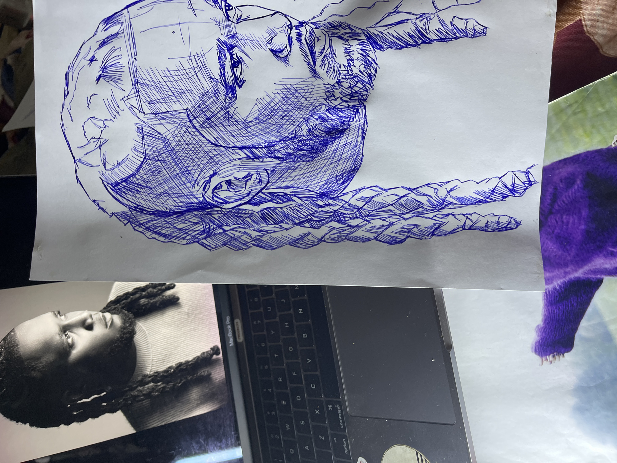 A blue ink sketch of a man's head and shoulders is shown. The sketch is rendered in a cross-hatching style, creating shadows and form. The man has braided hair that extends down to his shoulders and a beard. He is looking off to the side with his eyes slightly narrowed. The sketch is placed on a white piece of paper. To the left of the sketch, a portion of a laptop is visible, showing a black keyboard and trackpad. On the laptop screen, a black and white photograph of the same man is displayed, matching the subject of the sketch. The photograph shows the man wearing a light-colored sweater. Below the sketch, another photograph is partially visible, featuring a person wearing a bright purple sweater. The overall scene appears to be an artist's workspace, possibly in a home or studio. The image does not provide any information about the city of Jos, Nigeria.