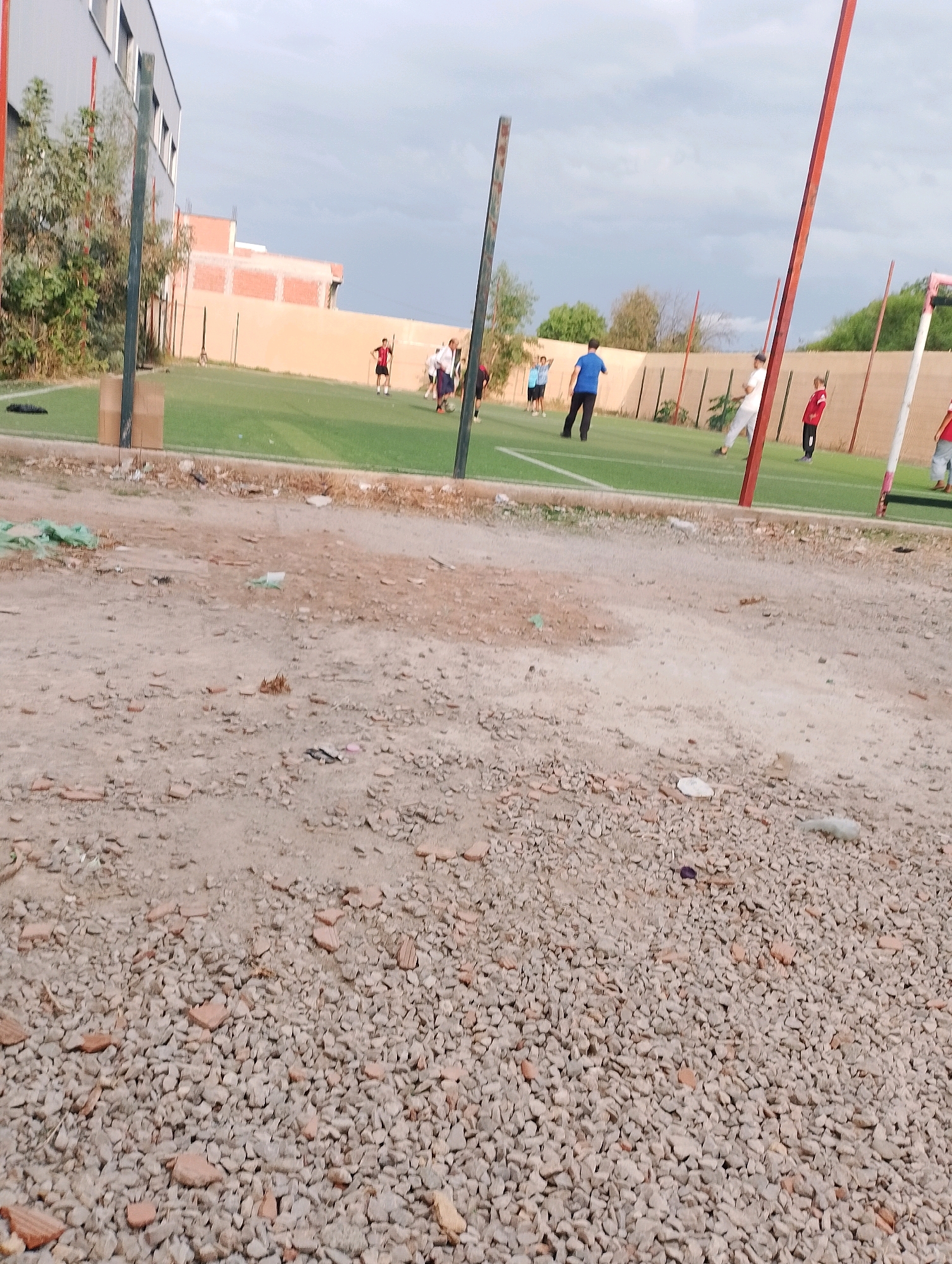 In Bordj Menaiel, Algeria, a group of individuals are engaged in a football match on an artificial turf field, bordered by a fence and a building on the left. The foreground is a rough, gravelly dirt area with scattered debris, suggesting an unfinished or unmaintained space adjacent to the sports facility. The sky is overcast.