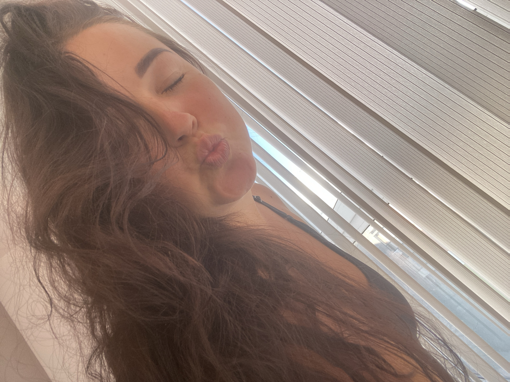 A young woman with long, dark, wavy hair leans her head towards the camera, puckering her lips. She has her eyes closed, and her eyebrows are slightly arched. Her skin appears flushed, and she is wearing a dark-colored top. The background is filled with the angled lines of white blinds, suggesting she is indoors near a window. Sunlight streams through the blinds, casting a bright, dappled light. The overall impression is of a casual, sun-drenched moment, possibly taken in the late morning or early afternoon given the brightness. There is no visible text in the image. The context of Sochi, Russia, implies a warm climate.