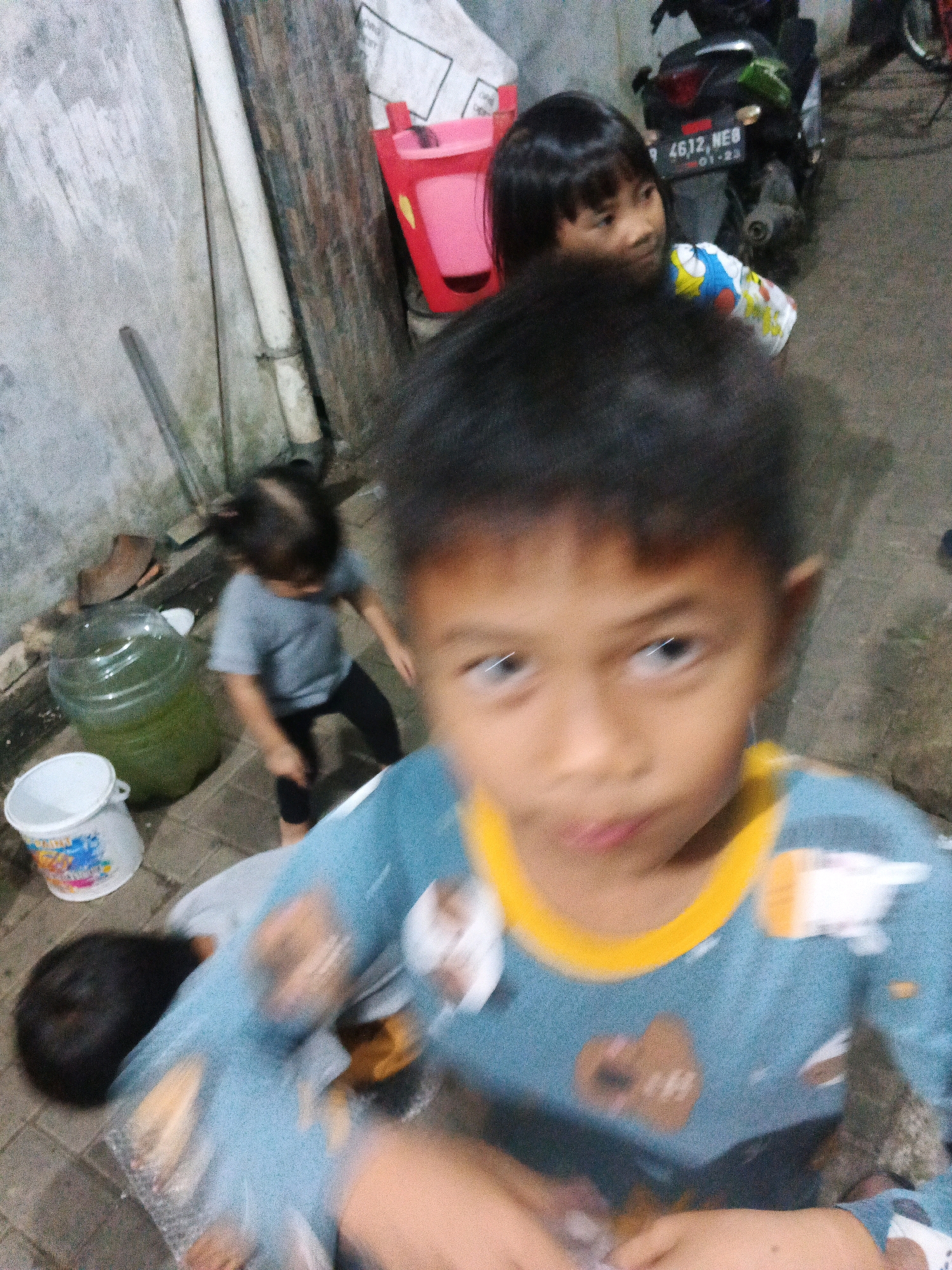 The image depicts a group of children in an outdoor urban setting. In the foreground, a child wearing a blue and yellow pajama set with cartoon characters is looking towards the viewer with a blurred expression. To the left and slightly behind this child, another child in a gray t-shirt and black pants is visible, appearing to be playing or moving. In the background, another child with dark hair is partially visible, wearing a shirt with a cartoon design. Further in the background, a motorcycle with a license plate reading "4612, NEB" is parked. Various objects are scattered around, including a large green plastic water jug, a white bucket with a cartoon print, and a red plastic container. The environment appears to be a paved outdoor area next to a textured wall. The scene is likely located in Kadu, Indonesia, given the style of the motorcycle license plate and the general surroundings.