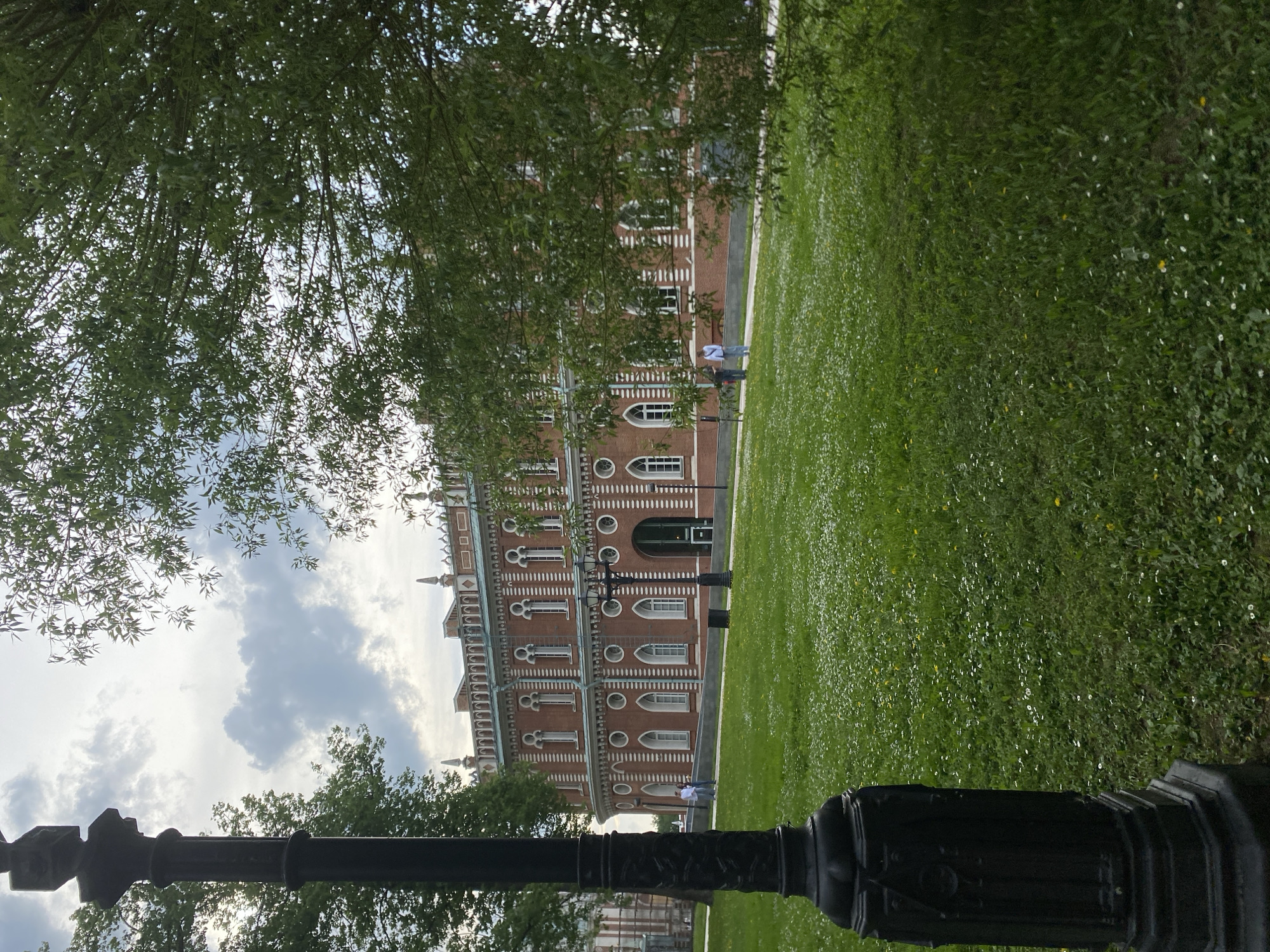 The image shows a large, red brick building with white trim, likely a historic structure, viewed from a low angle through a tree. A green lawn stretches out in front of the building. A couple of people are walking along a paved path next to the lawn. A black lamppost is visible in the foreground, slightly obscuring the view. The sky is partly cloudy. The location is Unknown, Unknown.