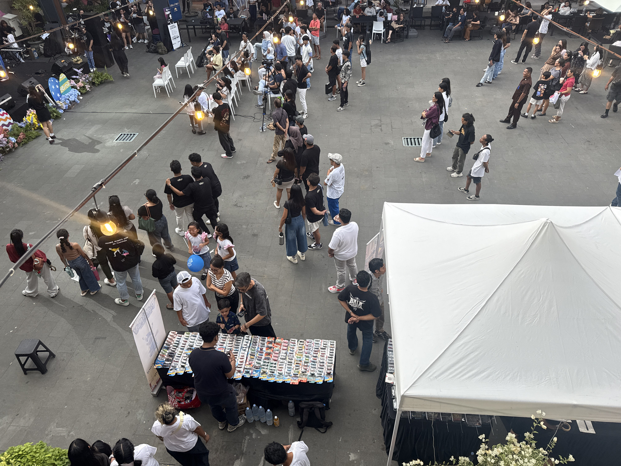 The image captures an elevated view of an outdoor event or market setting, likely in Kuta, Indonesia. The ground surface is a paved area, with a mix of light and shadows. A significant portion of the lower-right area is dominated by a large white tent structure with visible black drapery.
People are the main subjects of the image. The crowd comprises of individuals and small groups, of varied ages and ethnicities. Most people are dressed casually in clothing like t-shirts, jeans, shorts, and dresses, reflecting the warm climate.
Many appear to be attending a street fair or market, congregating around a table covered with merchandise, possibly toy cars or collectibles. Several people are seen interacting with the vendor and examining the items.
There are a few people sitting, some watching a stage that has floral decorations.
Lighting appears to be provided by a string of light bulbs suspended above the event space. Other objects present include chairs, tables, and a black stool. The overall scene suggests a lively public gathering or a weekend market.