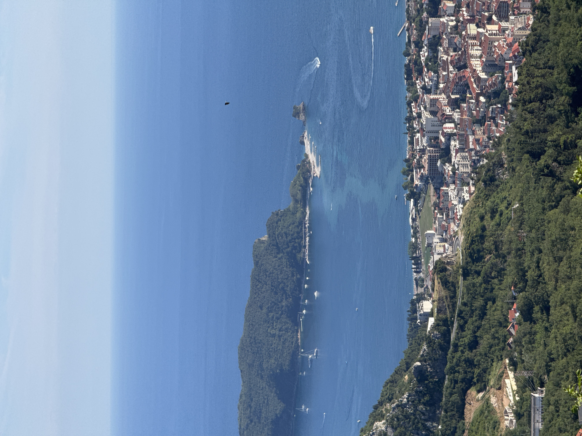 An elevated, wide-angle shot captures a coastal city nestled against a backdrop of a vast, blue ocean under a clear sky. The city, characterized by its densely packed buildings with red-tiled roofs and white facades, sprawls across the landscape. A winding road snakes through the green, tree-covered hills that flank the city. In the deep blue water, several sailboats are anchored, and a speed boat leaves a white wake. A lone bird is visible flying in the sky above the water. Further out, a large, green, tree-covered island juts out into the sea, with a smaller, sandy island and rocky outcrops situated near its coast. The horizon line is clearly defined, separating the water from the pale blue sky.