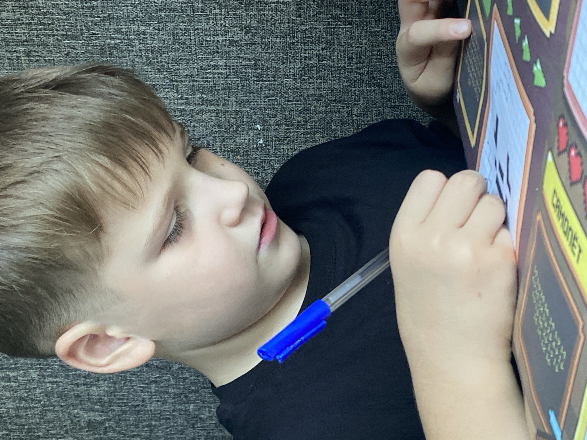 A young boy, with light brown hair and fair skin, is shown from the chest up, leaning over a table and concentrating on a piece of paper. He is wearing a black t-shirt and is holding a blue pen in his right hand, actively writing or drawing. His left hand rests on the table, near the paper, with his fingers slightly bent. The paper appears to be part of a workbook or activity sheet with grids and colorful illustrations. The background is a dark, textured surface, possibly a wall. The lighting is soft, highlighting the boy's face and hands. No specific location within Chaikovsky, Russia, can be determined from the image alone.