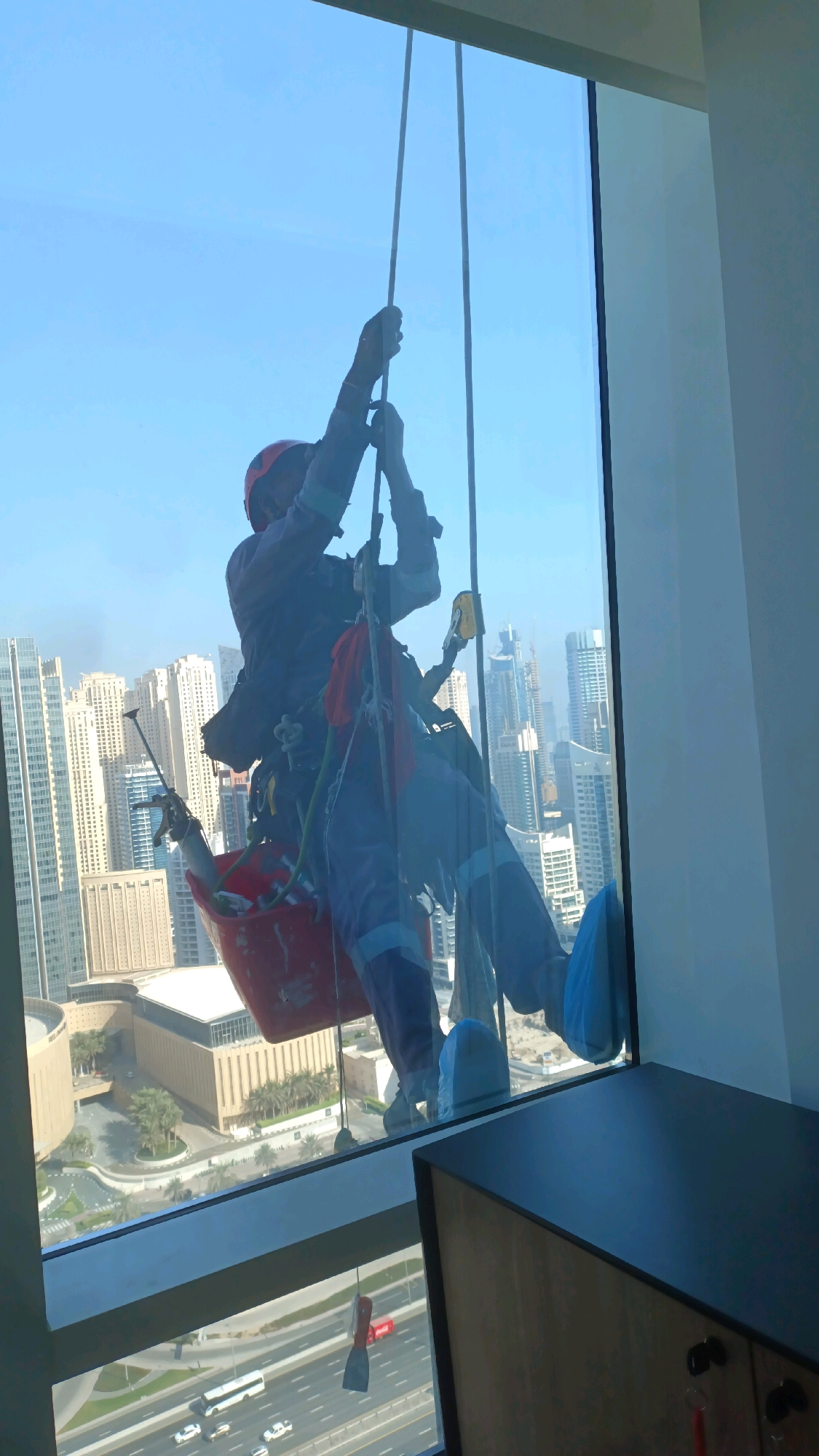 A window cleaner, suspended by ropes on the exterior of a skyscraper, cleans the glass in Dubai. The cleaner wears a red helmet, dark blue overalls, and protective blue shoe covers. They are secured by a harness and ropes attached to a red bucket containing cleaning supplies. In the background, the cityscape of Dubai is visible, with numerous skyscrapers, including the Princess Tower and 23 Marina Tower, a highway with cars and a bus, and a modern building complex. The scene takes place during the daytime under a clear blue sky.