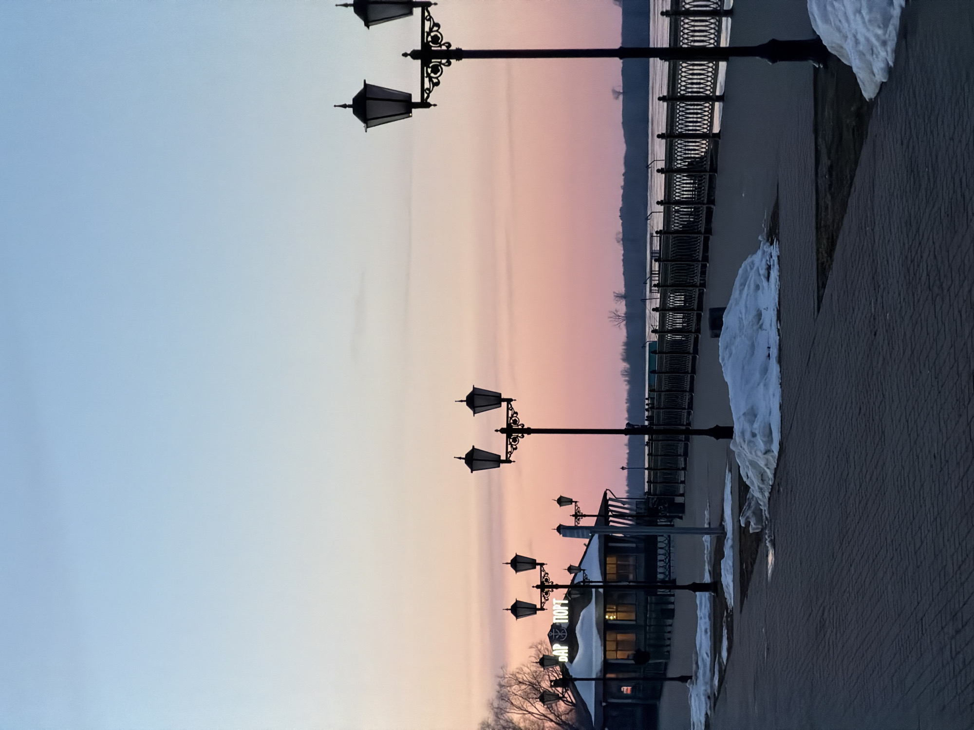 The image depicts a waterfront promenade in Sarapul, Russia, at dusk or dawn. The sky transitions from blue to pink above a body of water with a treeline visible on the opposite bank. Along the promenade, a row of ornate streetlights is visible, casting reflections on the wet pavement. Patches of snow remain along the edge of the walkway. A building, possibly a restaurant or bar labeled "БАР ПОРТ," is partially visible. A metal railing separates the walkway from the water.
