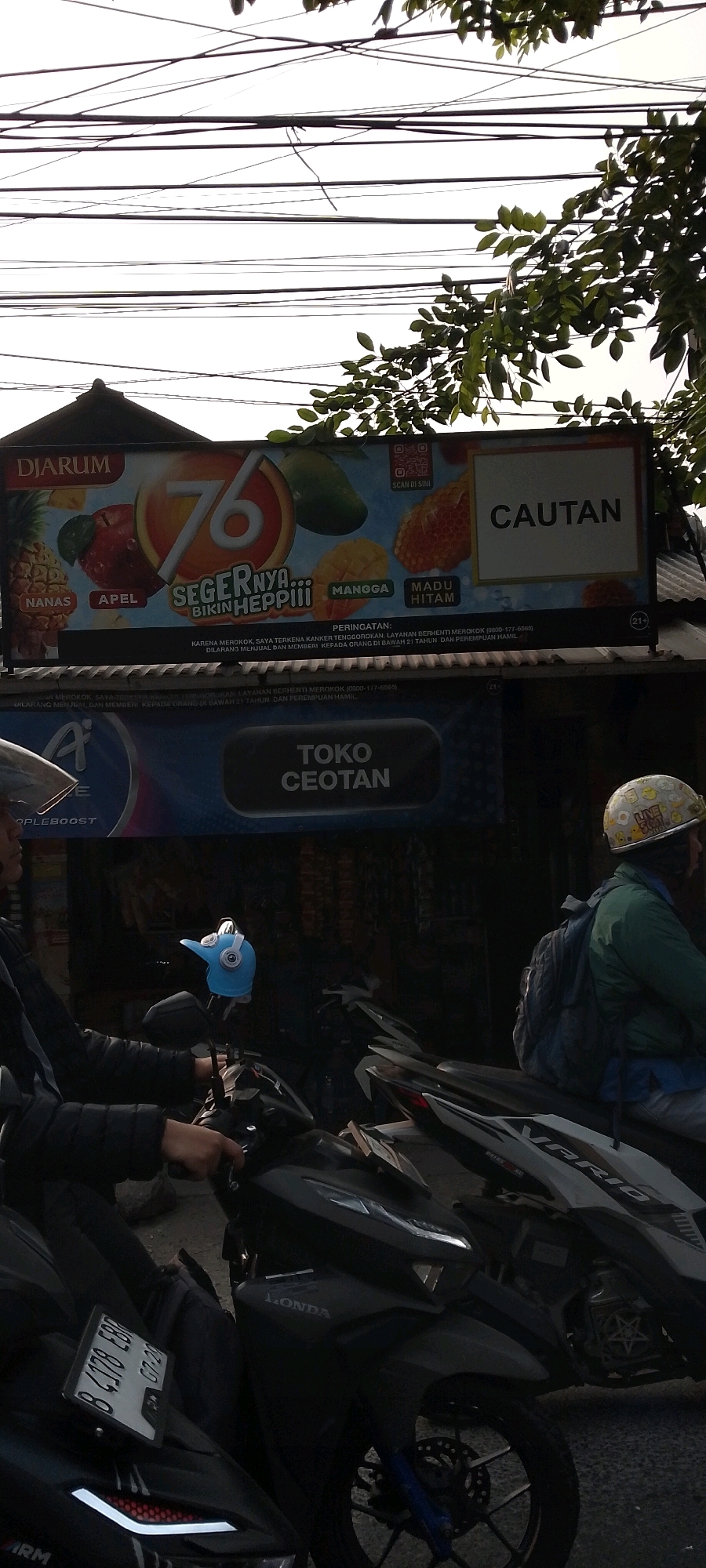 The image depicts a street scene in Bekasi, Indonesia, under an overcast sky. Overhead, a tangled mess of electrical wires crisscrosses the frame, partially obscured by lush green tree branches. Dominating the upper portion of the image is a large billboard for "Djarum 76," featuring colorful depictions of fruits like pineapples, apples, mangoes, and jackfruit. Below the billboard, a shop sign reads "TOKO CEOTAN."
In the foreground, several motorbikes are parked. A person wearing a black jacket and a blue helmet with headphones is seated on one of the motorbikes, holding the handlebars. Another person, wearing a green jacket and a helmet adorned with cartoonish stickers, is also on a motorbike, with a backpack slung over their shoulder. The motorbikes are dark-colored, with visible details like headlights, license plates, and wheels. The overall impression is a busy urban environment, likely a commercial area, with a focus on street-level commerce and transportation.
