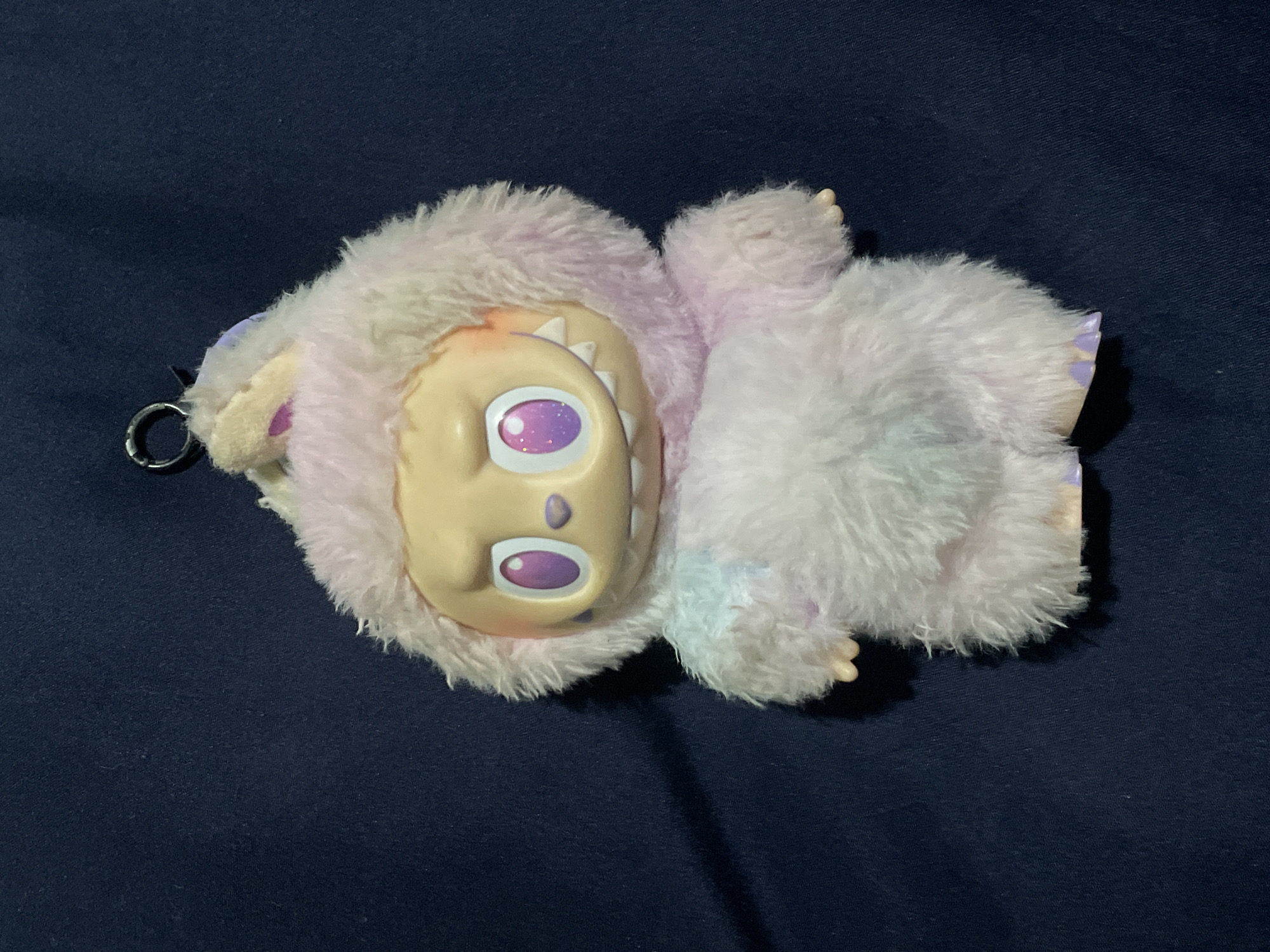 A plush toy, resembling a mythical creature or monster, is lying face-up on a dark navy or indigo fabric surface. The toy features a light peach-colored face with large, round purple eyes that contain sparkling, glittery specks. Small white teeth are visible inside what appears to be its open mouth or a distinct facial feature. The toy's body is covered in soft, fluffy fur that transitions between pastel shades of white, light pink, and light blue/purple. Small, peach-toned hands and feet, which resemble tiny claws or paws, protrude slightly from the fur on its limbs. A metallic silver keyring is attached to the toy's left side. The lighting is somewhat dim, casting soft shadows around the toy and highlighting its textures. The overall impression is that of a cute, fantastical character. There is no visible text in the image.
