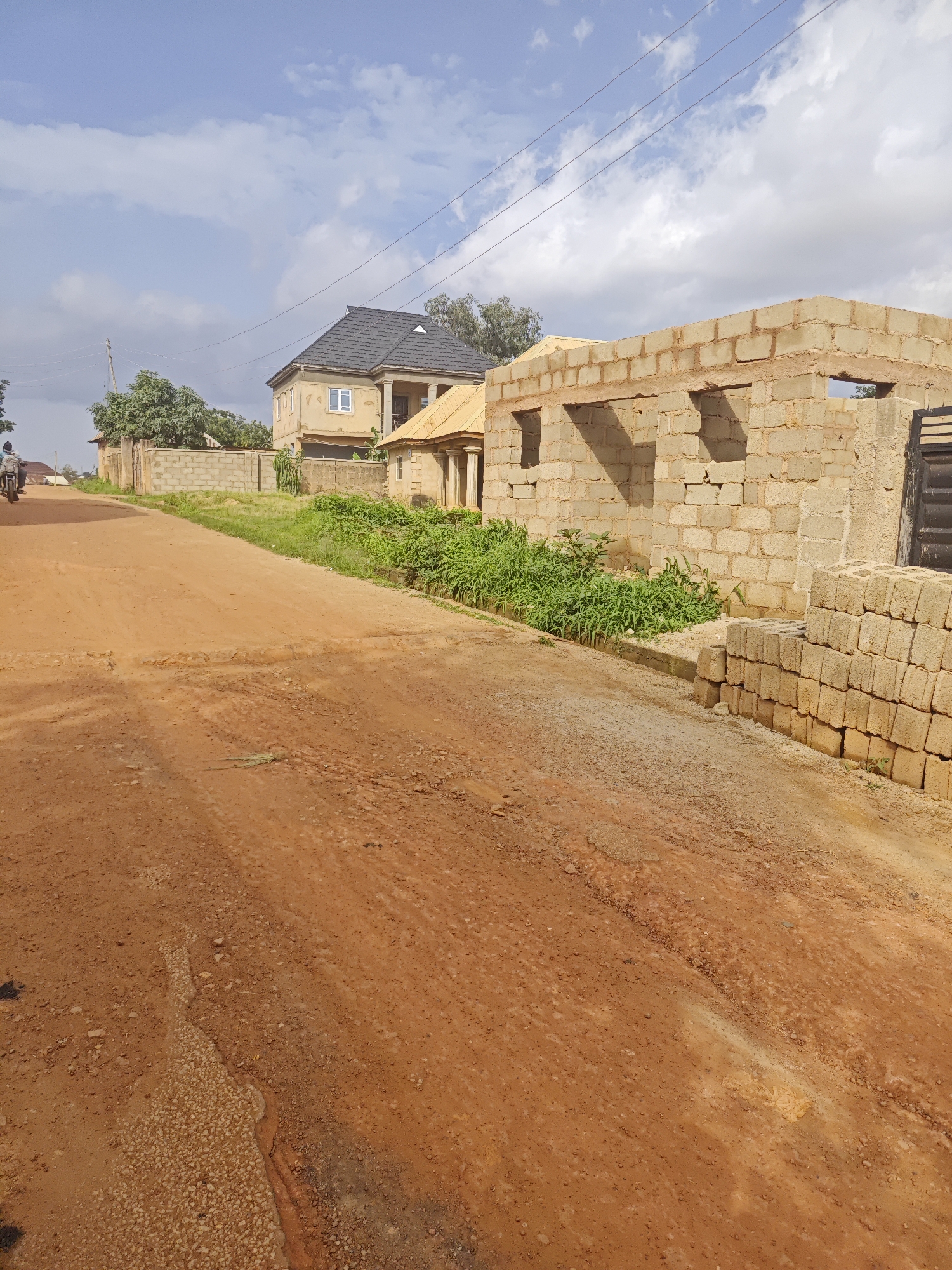 A clear, slightly overcast day in Bukuru, Nigeria, features a dirt road leading uphill past residential construction. The road is predominantly red soil, with some gravelly patches and visible tire tracks. On the right side of the road, a partially constructed building made of cinder blocks stands, with stacked blocks to its right. A finished house with a dark grey roof and beige walls is visible further up the road on the left, with a smaller building with a yellow roof beside it. Green grass and foliage grow along the sides of the road and around the buildings. A person is seen riding a motorcycle uphill on the left side of the road. Power lines are strung across the sky.