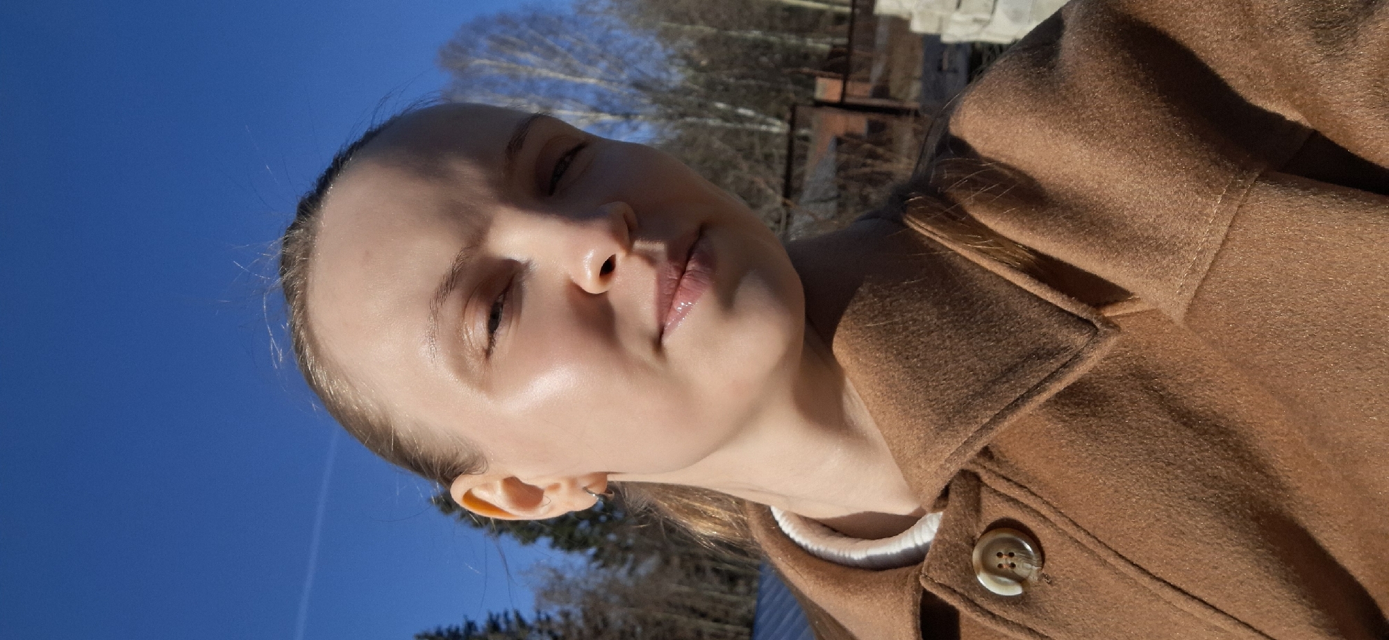 A young woman with fair skin and blonde hair poses outdoors in Chaikovsky, Russia. She wears a brown wool coat with a white collar and has her eyes closed. The sky is blue, and trees are visible in the background. There is also a glimpse of an industrial or construction area behind her.