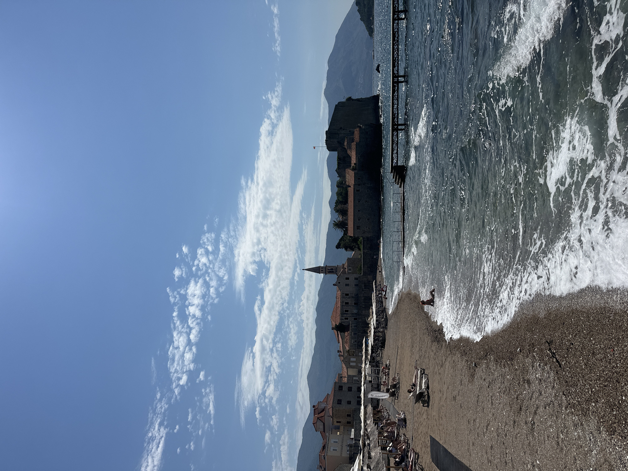 This is a medium shot of a sunny day at a beach in Budva, Montenegro. The beach is made of small pebbles and sand, with numerous lounge chairs and umbrellas set up. Several people are visible on the beach, some lounging, some sitting. A single person is sitting in the shallow water near the shore. The water is a deep blue and choppy with whitecaps. In the background, there is a historic town with red-tiled roofs and a prominent church steeple, situated on a rocky peninsula. A stone fortress is visible on the end of the peninsula. Mountains form the backdrop, and a wooden pier extends into the sea. The sky is a clear blue with scattered white clouds.