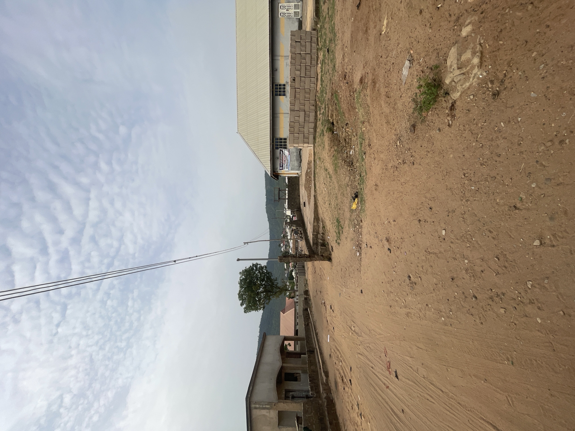 The image captures an outdoor scene depicting a semi-urban or industrial area under an overcast or partly cloudy sky. The perspective is from ground level, looking towards distant hills.
The foreground is dominated by a wide, unpaved dirt road or path, covered in light brown earth, small stones, and faint tire tracks. Scattered debris, including pieces of litter, is visible on the ground.
On the right side of the frame, a large industrial-style building stands prominently. It features a light-colored, possibly corrugated metal roof and grey walls with multiple windows. Outside this building, large stacks of grey concrete blocks are neatly arranged. A blue and white banner is affixed to the building's facade, bearing partially legible text which includes "ENTERPRISE", "GROUP", and possibly "NIGERIA".
On the left side, closer to the viewer, there is a smaller, seemingly unfinished residential building with unpainted cement walls and a concrete porch area. A single large tree with green foliage is situated near the center of the image, behind this smaller building, and partially obscures the view of other structures in the mid-ground.
Overhead, three parallel power lines stretch across the upper portion of the sky, connected to utility poles which are not fully visible. The sky shows distinct patterns of cirrus or altocumulus clouds.
No people are visible, and no specific activity is taking place, presenting a static view of the environment. The time of day appears to be daytime, under bright but diffused light. The setting is consistent with the provided context of Lokoja, Nigeria.