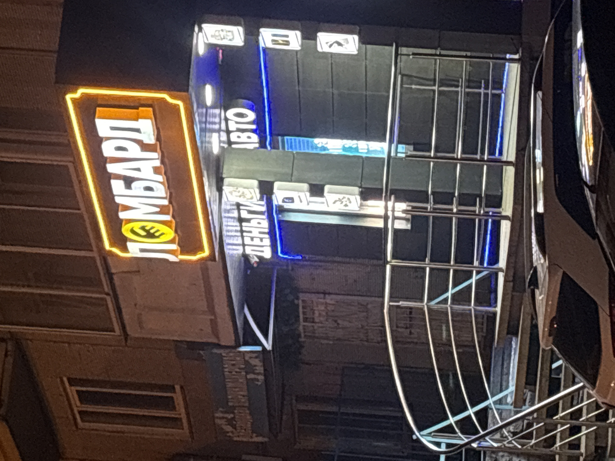 The image shows the facade of a building in Krasnoyarsk, Russia. A brightly lit sign for a "Lombard" (pawn shop) with a yellow border is visible. Next to it is a blue illuminated sign with "ABTO" (AUTO) and "ДЕНЬГИ" (MONEY) in Russian. The facade includes metal railings and windows. A white car is partially visible on the right side of the image. It is night.