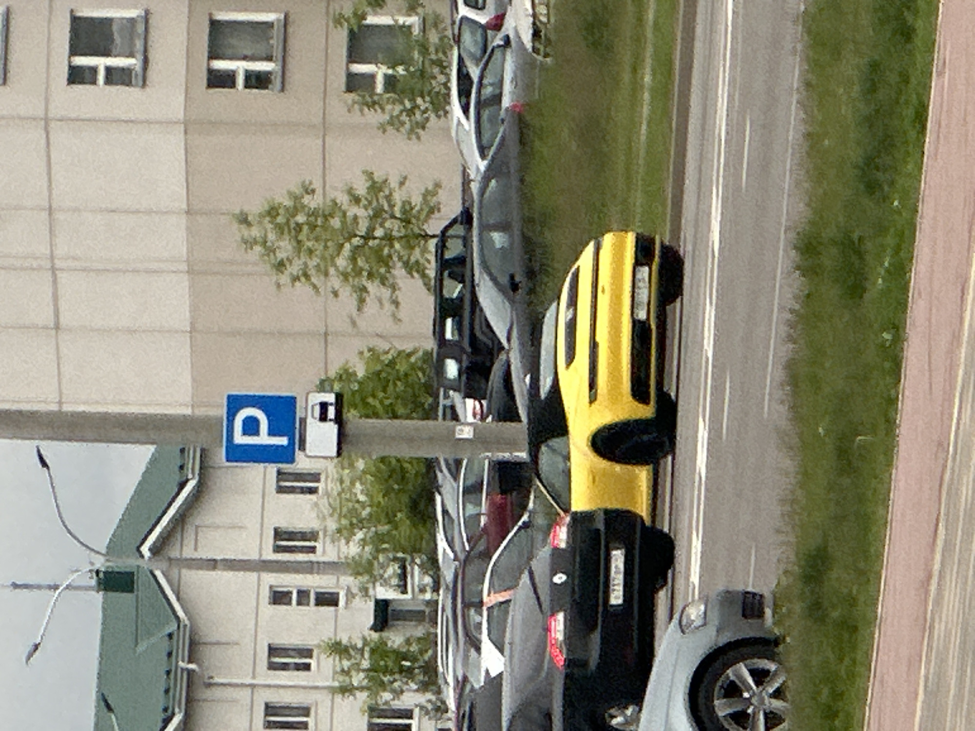 The image, taken in Бараўлянскі сельскі Савет, Belarus, features a street lined with parked cars adjacent to a multi-story building. A yellow car is prominently positioned on the street, followed by a black car, and then a gray car. A parking sign with a "P" is visible next to the building. The building facade has windows. A tree stands between the building and the cars. A road with lane markings runs alongside the parked cars. A sidewalk is on the right side of the image.