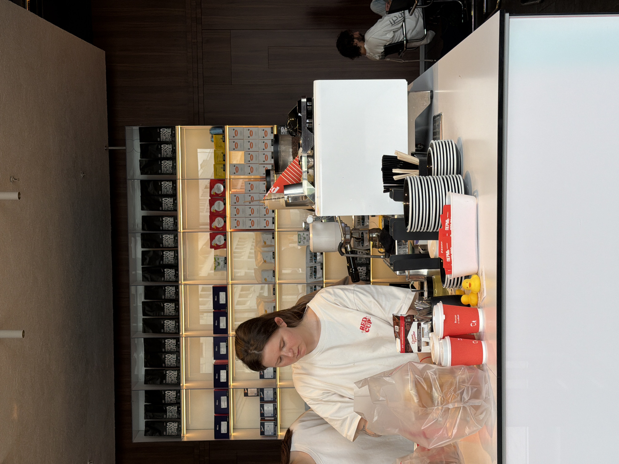 A young woman with brown hair, wearing a white t-shirt with a red logo that reads "RED CUP", stands behind a white counter in what appears to be a coffee shop. She is focused on packaging items in a clear plastic bag. On the counter, there are two red coffee cups, two yellow rubber ducks, a pink container, a box of coffee, a stack of black and white striped plates, and various coffee-making equipment. In the background, shelves are stocked with bags of coffee and boxes. To the right, a man with dark curly hair wearing a light-colored sweatshirt sits in a chair, looking towards the counter. The environment suggests a modern coffee shop setting. Given the presence of Russian text on some of the packaging and the man's attire, the location is likely Perm, Russia.