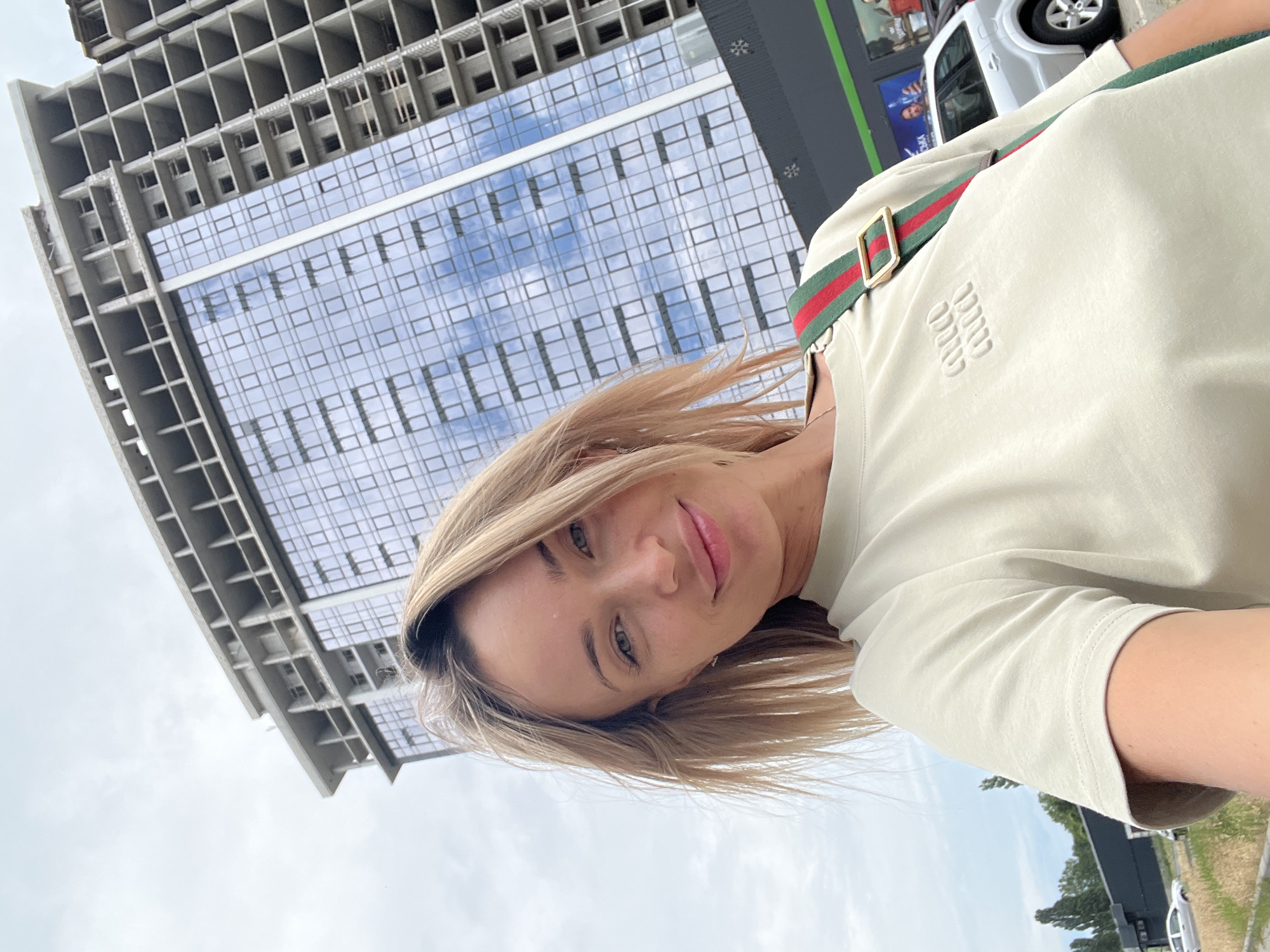 A young Caucasian woman with blonde hair and green eyes is taking a selfie outdoors. She is wearing a light-colored t-shirt with a logo and a strap from a bag with green and red stripes. Behind her, a tall, unfinished concrete building with many windows is visible, reflecting a cloudy sky. To the right of the building, a silver car is parked. The scene appears to be in an urban environment, possibly Fontanka, Ukraine. The overall lighting suggests it is daytime.