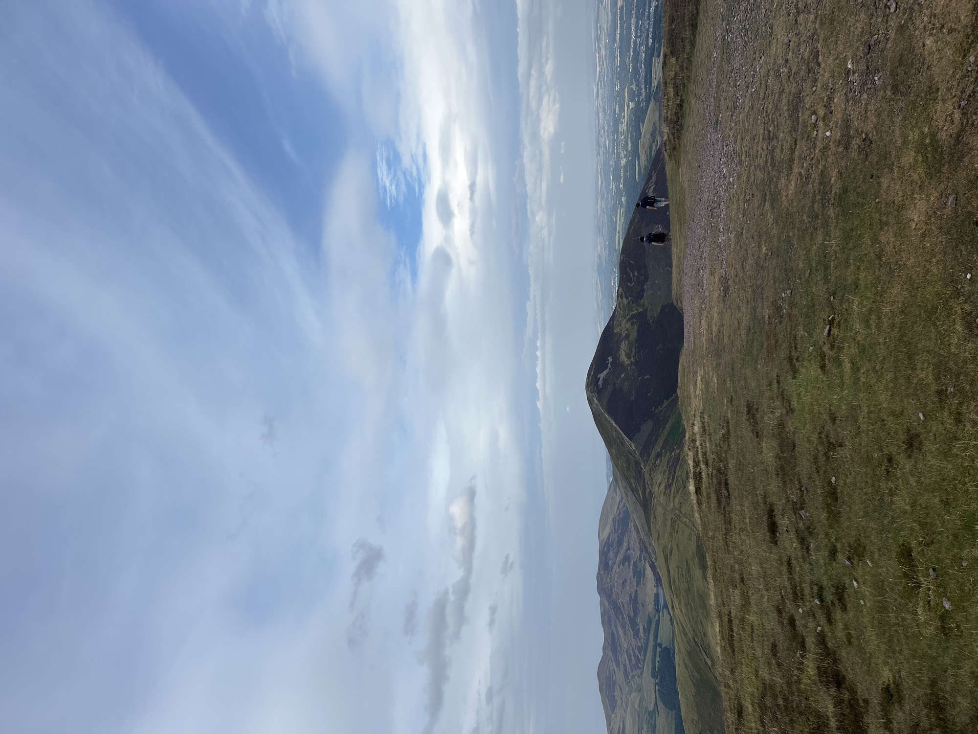 Two individuals stand on a grassy hilltop overlooking a valley with rolling hills and a distant town. The sky above is filled with a mix of white and grey clouds. The foreground is dominated by a rough, grassy slope with scattered rocks. In the mid-ground, a conical hill with dark vegetation rises prominently. Beyond this, a panoramic view of a landscape unfolds, showing further hills, fields, and a cluster of buildings that form part of the town of Penicuik. The two people appear to be hikers, facing away from the camera and towards the vista. The overall scene depicts a natural landscape with human presence, suggesting an outdoor recreational setting near Penicuik, United Kingdom.