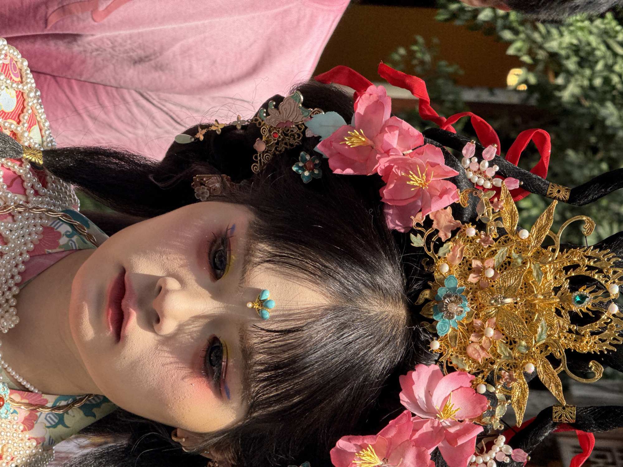 A close-up, low-angle shot of a person in traditional Chinese attire. The person has pale skin, black hair adorned with ornate gold and floral hairpieces, and wears heavy eye makeup with shades of pink and yellow. A turquoise ornament is affixed to their forehead. The person is wearing a garment with a pearl-beaded collar. The backdrop includes a blurred view of greenery and architectural details, possibly indicating an outdoor setting in the Yuzhong District, China.