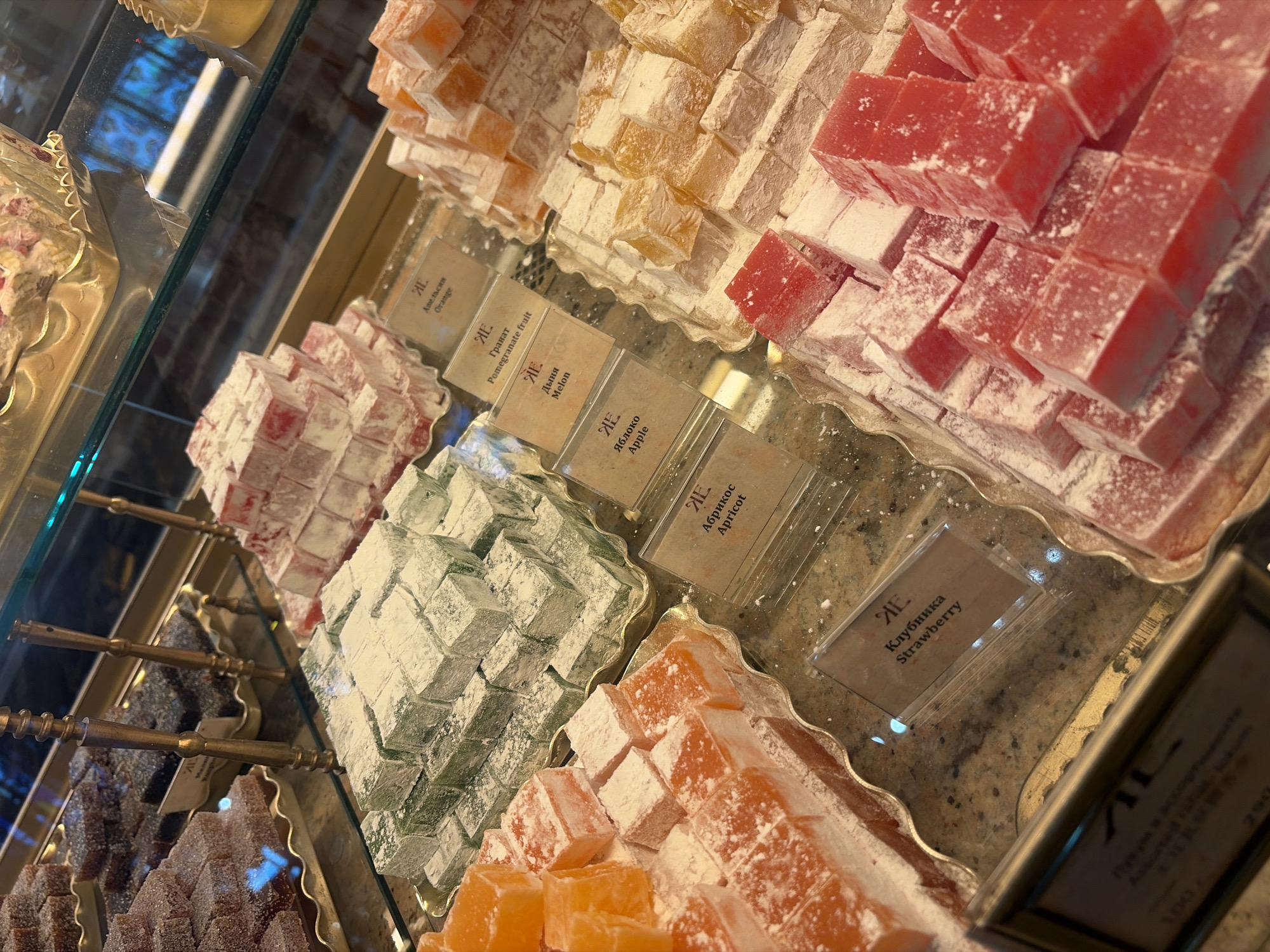 The image shows a display of Turkish delight in a confectionery shop, likely located in Saint Petersburg, Russia. The Turkish delight is arranged in tiered piles on ornate gold-colored trays, covered in a dusting of powdered sugar. Various flavors are visible, including orange, green (possibly pistachio or mint), yellow (possibly lemon or apricot), and red (likely strawberry or rose). Small labels identifying the flavors are placed in front of some of the piles, with text in both Russian and English, such as "Orange," "Pomegranate fruit," "Melon," "Apple," "Apricot," and "Strawberry." The display is enclosed in a glass case, reflecting the interior lighting and the surrounding environment, which appears to be a well-lit shop. The overall impression is one of a high-end dessert shop, showcasing a variety of colorful and inviting sweets.