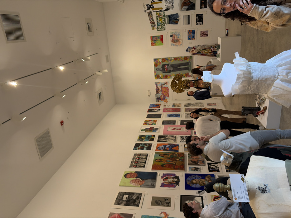 The image depicts an art exhibition or gallery opening within the Wynwood Arts District in Miami, Florida, United States of America, specifically indicated by signage mentioning "Wynwood Walls" and "Miami-Dade County Public Schools." The environment is a brightly lit, white-walled room with a polished concrete floor and a high ceiling equipped with linear track lighting and rectangular ventilation grates. A red fire alarm sensor is visible on the ceiling.
Approximately 10-15 individuals, primarily adults, are present throughout the room, engaging with the art and each other. Most individuals are dressed in casual to business-casual attire. On the left side of the frame, two men are conversing near a display bust. In the center, a man with dark hair in a light-colored shirt is talking, facing left, with his hand near his mouth. Another man in a white long-sleeved shirt stands behind him, facing away from the camera. Towards the middle-right, a group of people is gathered, including a woman with long dark hair looking left, and a man in a dark suit standing near an ornate golden headdress displayed on a stand. In the foreground on the right, a woman with glasses and curly dark hair is looking towards the right, with her hand near her chin.
The white walls are extensively covered with a diverse collection of artworks, including paintings, drawings, and mixed media pieces, displayed from near floor level to high on the walls. Styles range from realistic portraits, such as an elderly couple on the left wall and a family scene in the middle-left, to abstract and pop art designs. Prominent artworks include a large painting of a figure against a floral background on the right wall, and several pieces incorporating text and fashion elements, such as denim shorts and a piece featuring the text "BLAAH BLAAH."
Several sculptural or fashion displays are positioned on white pedestals across the room. In the immediate foreground on the left, a headless mannequin bust, likely used for clothing display, is visible with a white information card attached. In the foreground on the right, a white, ruffled, sleeveless dress is displayed on another headless mannequin bust. Further into the room, on a white pedestal, a black boot, a red high-heeled shoe, and a metallic or crumpled paper-like sculpture are exhibited. Adjacent to these, a golden, elaborately adorned headdress is displayed on a bust.