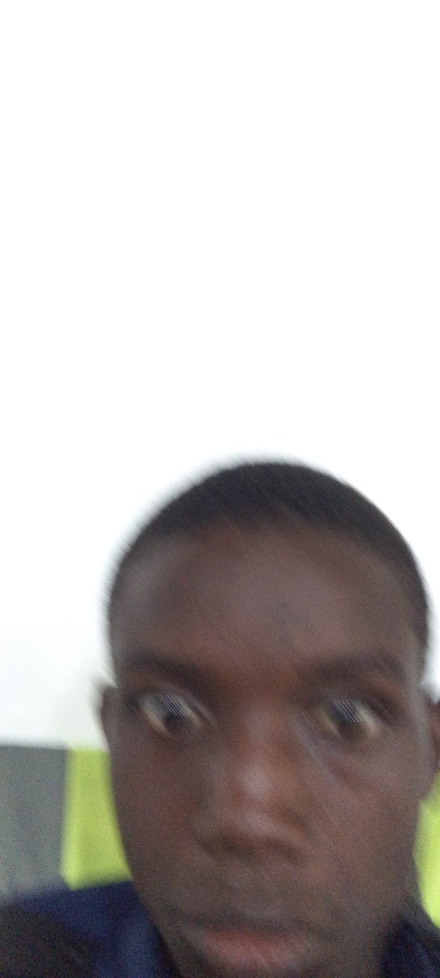 A blurry close-up selfie of a dark-skinned person with short hair. The background is mostly white, with a slight green and gray area visible in the lower left.