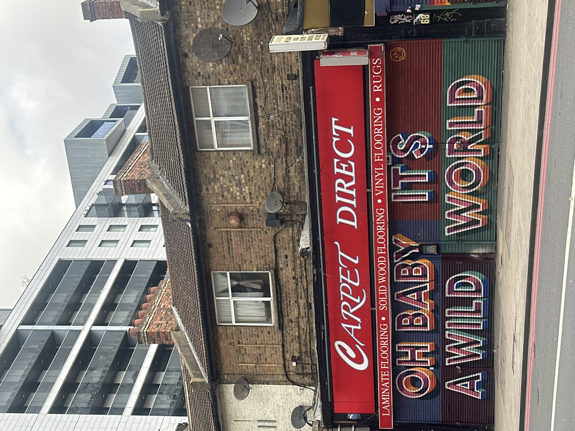 The image depicts a building facade in London, United Kingdom, with a "Carpet Direct" storefront prominently displayed. The storefront features a red sign with white lettering advertising laminate, solid wood, and vinyl flooring, as well as rugs. Adjacent to it is a metal security shutter painted with the phrase "OH BABY IT'S A WILD WORLD" in vibrant, neon-style lettering.
The building itself is constructed of brick, with multiple windows, satellite dishes, and visible wiring. A modern, multi-story glass and steel building rises in the background, contrasting with the older architecture of the storefront. The street is partially visible in the foreground, marked with a red line. The sky is overcast, creating soft lighting.