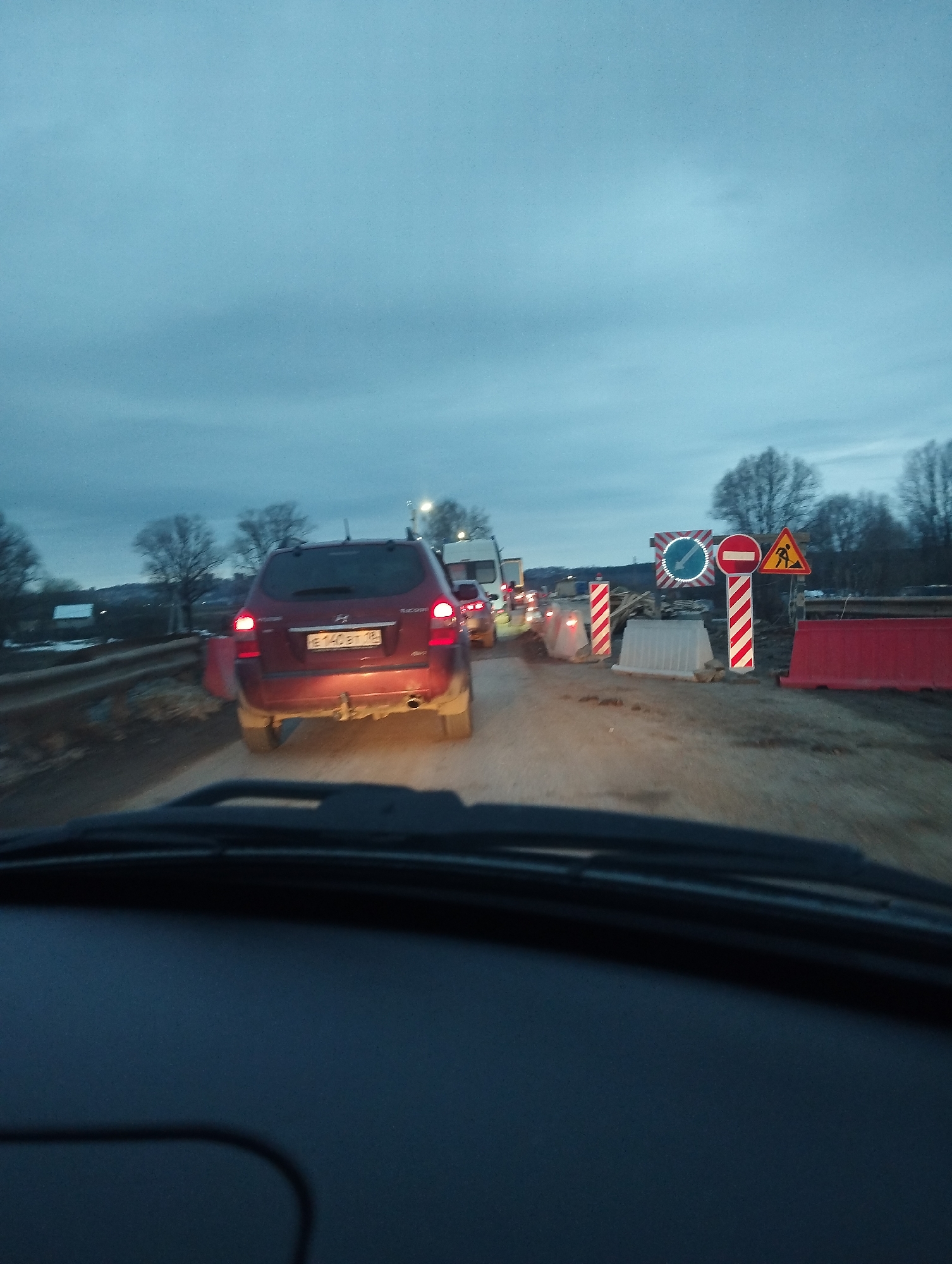 The image shows a view from inside a car, looking out towards a road with traffic and construction work in progress. A red SUV is directly in front of the viewer's car. Further ahead, several vehicles are visible, possibly waiting in a queue. Road work barriers and signs, including a "no entry" sign and a "road work" sign, mark the construction zone. The scene appears to be in Gavrilovka, Russia, as indicated by the car registration plate on the red SUV. The sky is overcast. Trees are visible in the background on either side of the road.