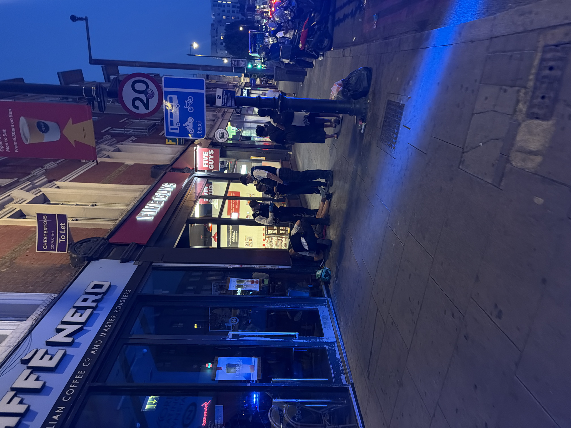 The image shows a street scene in London, United Kingdom, at dusk. In the foreground, there is a "Caffe Nero" storefront, identified by its blue and white signage: "ITALIAN COFFEE CO AND MASTER ROASTERS". A "To Let" sign from "CHESTERTONS" is visible above the cafe. Next to it is the storefront of "Five Guys". Several people are standing or kneeling in front of Five Guys. A blue sign indicates a taxi and motorcycle route. A "20" speed limit sign is also visible on the post. In the background, more shops and streetlights can be seen. Bags of refuse are on the sidewalk.
