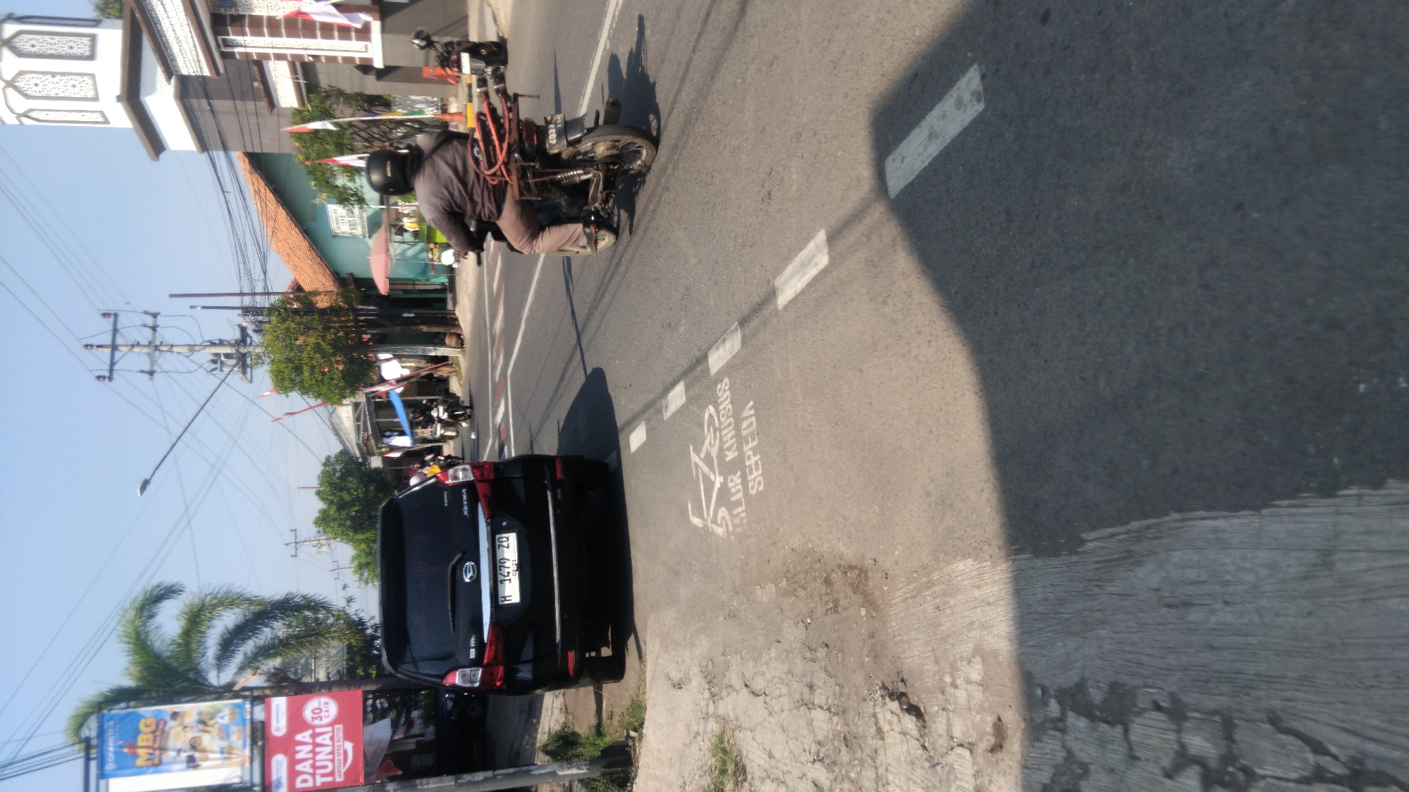 The image depicts a street scene in Proyonanggan Tengah, Indonesia, under a clear blue sky. On the left, a black SUV with the license plate "H 1479 ZQ" is parked on the side of the road. Adjacent to the SUV is a sidewalk with a clear marking indicating a "BIKE LANE" with a bicycle symbol and the text "JALUR KHUSUS SEPEDA". Further down the street, a person is riding a motorcycle with a red rack on the back, wearing a helmet and brown pants. The motorcycle has the number "4037" visible on its license plate. Several other motorcycles are also visible further down the road, suggesting a moderately trafficked area. Buildings with traditional Indonesian architecture, including a mosque with a minaret, line the right side of the street. Small shops and trees are also present. The road surface is asphalt, and there are visible white lane markings. The overall impression is that of a typical urban street in Indonesia.