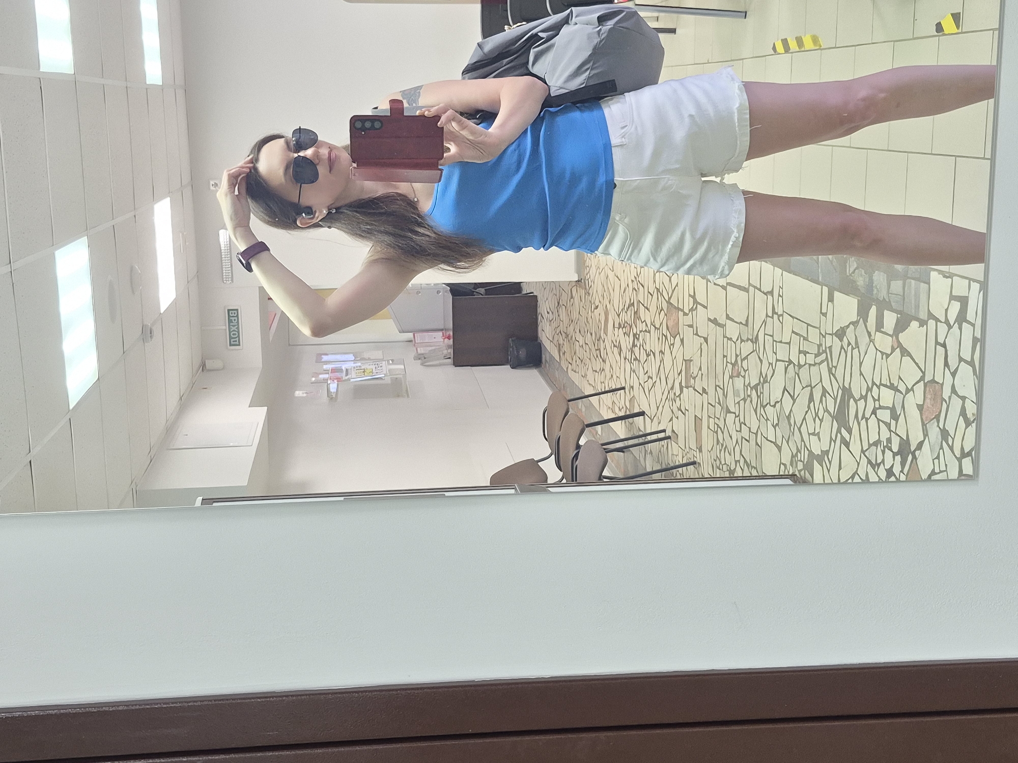 A mirrored image shows a woman in a blue tank top and white shorts taking a selfie. She is wearing sunglasses and a watch. A grey backpack is nearby. The background includes a tiled wall and floor, office furniture, and overhead lighting.