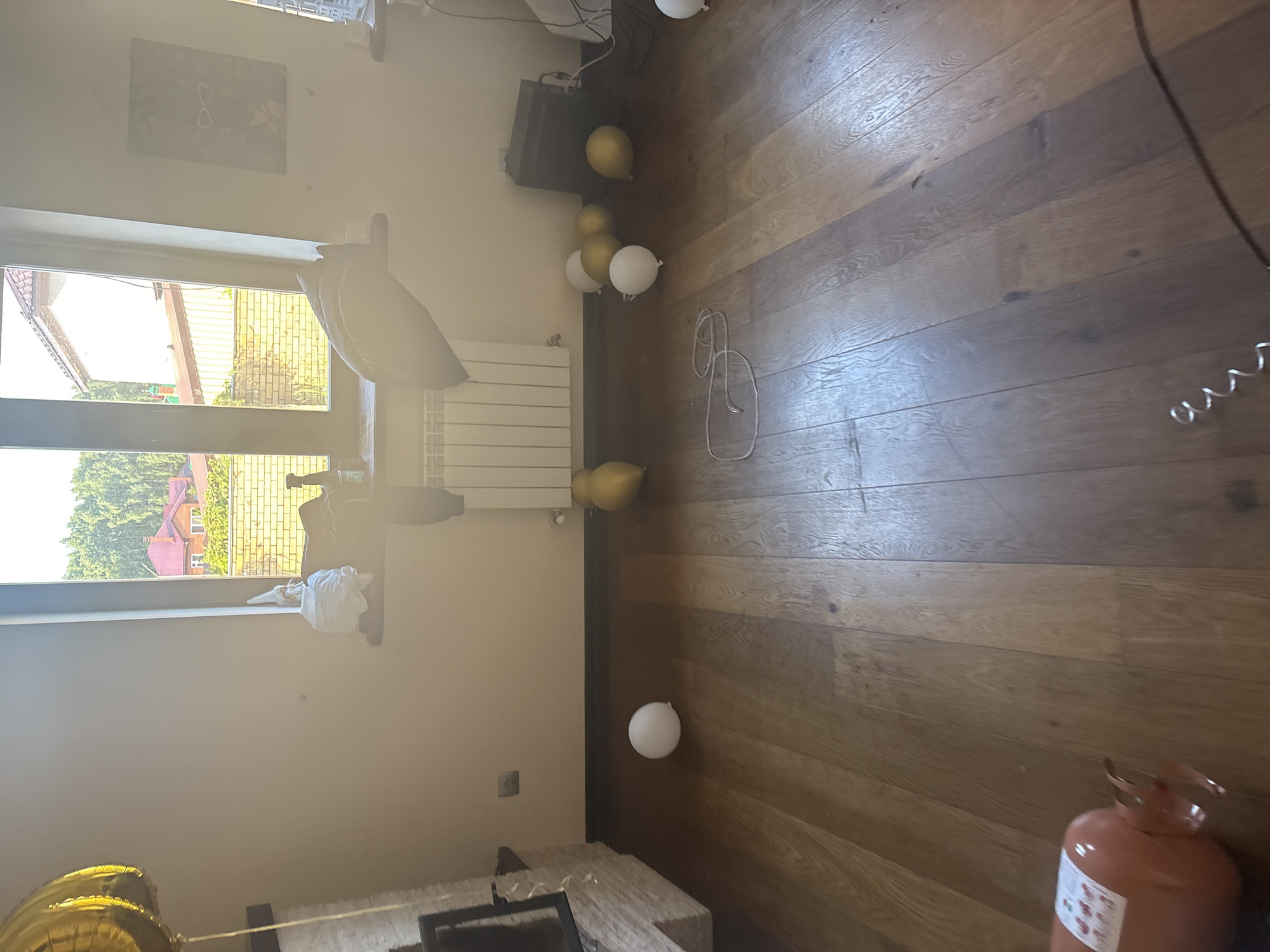 The image shows an indoor scene, possibly a living room, with wooden flooring. Several balloons, both white and gold, are scattered across the floor. One large gold balloon is partially visible in the upper left corner. A gas cylinder, orange in color, lies on its side in the lower left portion of the image. In the background, there is a window overlooking a residential area with houses and trees. A radiator is visible beneath the window, and a shelf with some objects is above it. There are also some electronic devices and cables on the floor in the right background. No people are present in the image. The scene appears to be located in the city of Orekhovka, Russia, as per the provided context.