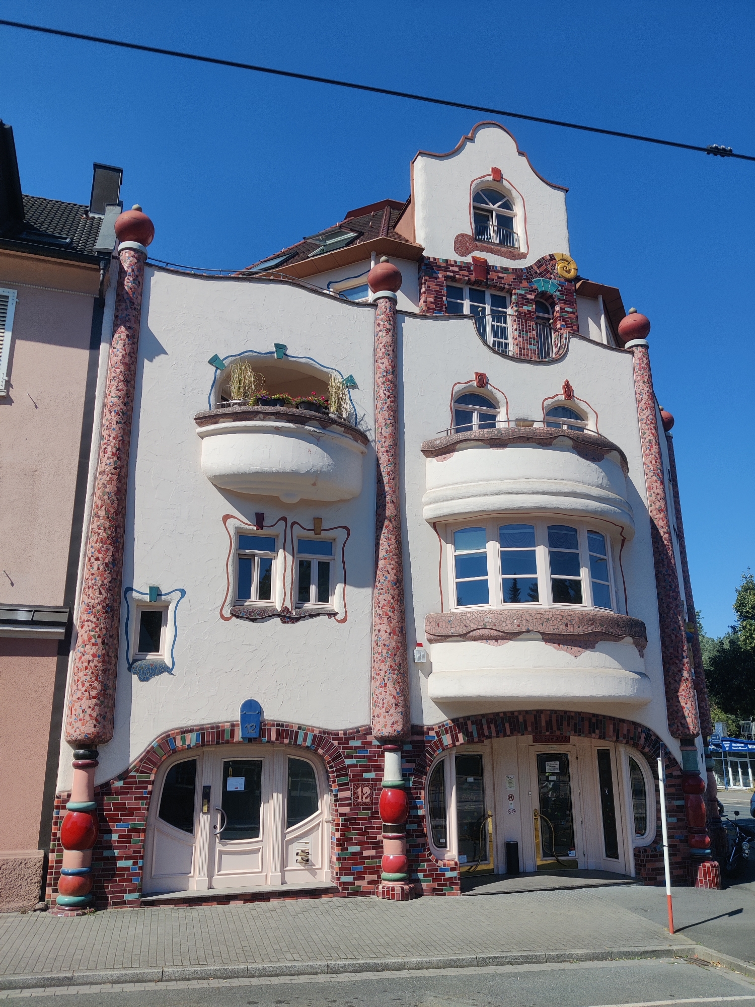 The image shows a brightly colored, unconventional building in Dortmund, Germany. The building's facade is a mix of white stucco, colorful mosaic tiles, and red brick. It features several curved balconies, rounded window frames, and whimsical decorative elements, including large, spherical finials topping the columns that flank the entrance. The entrance itself is an arched doorway with double doors, framed by colorful brickwork. Above the entrance, the building's design becomes more elaborate, with multiple levels of windows and balconies, culminating in a steeply pitched roof. The sky is a clear, bright blue, indicating fair weather. The building appears to be situated on a street with a sidewalk and what looks like a road in the foreground. There are no people visible in the image. The architectural style is reminiscent of Hundertwasser's designs, known for their organic forms and vibrant colors.