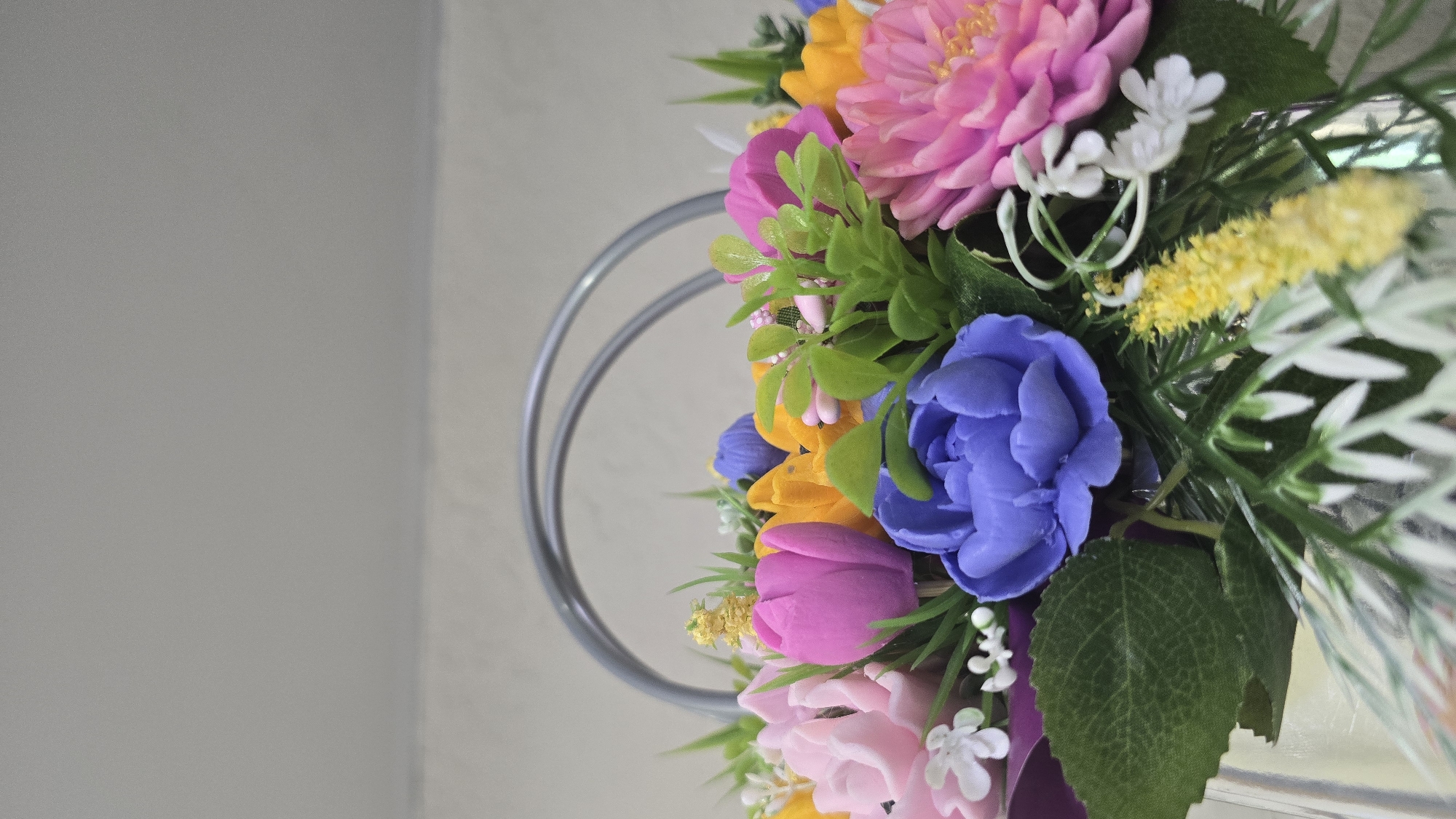 A close-up, eye-level shot depicts an arrangement of numerous vibrantly colored artificial flowers and foliage, positioned within a container with a visible silver-colored handle. The flowers appear to be crafted from a solid, malleable material, possibly soap. Prominent blooms include a large, multi-petaled blue flower in the central foreground, a bright pink tulip-like flower to its immediate left, and a lighter pink, multi-petaled flower further to the left. To the right, a large pink flower with distinct central stamens is visible. Yellow tulip-like flowers are interspersed throughout the arrangement, particularly behind the central pink tulip. Small white filler flowers, some with multiple delicate petals and others resembling tiny bells, are scattered among the main blooms. Thin, yellow, spiky floral elements are also present. The arrangement incorporates various shades of artificial green foliage, including broad leaves (e.g., on the lower left), delicate fronds, and small clusters of lighter green leaves. A metallic, silver-colored, arced handle, likely belonging to a basket or similar container, is visible above the flower arrangement. The background consists of a plain, light grey wall, with a subtle horizontal demarcation line separating a slightly lighter upper section from a marginally darker lower section. There is no discernible information within the image to indicate its specific geographic location in the city of Kazan, Russia, or any other location.