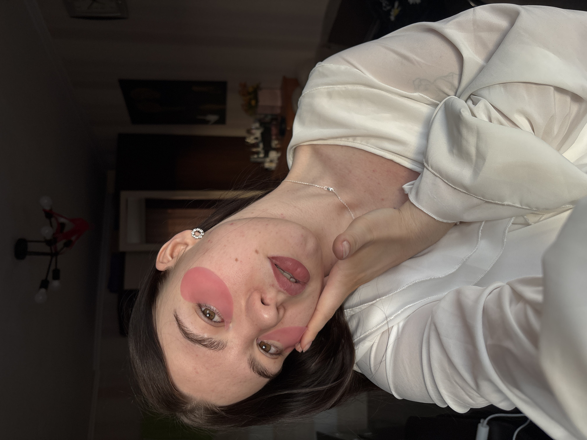 A woman is pictured indoors. She is wearing an off-white, silky blouse. She has pink patches under her eyes and on her cheek. Her right hand touches her face. A thin, silver necklace adorns her neck. There is a chandelier on the left and dark furniture is visible in the background. The image was taken in Perm, Russia.