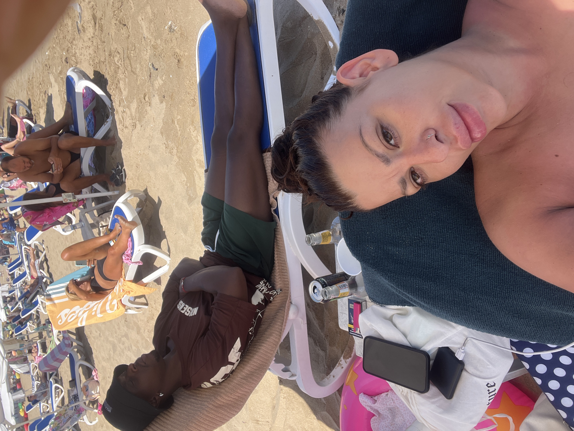 The image depicts a sunny day at a crowded beach in Sozopol, Bulgaria. In the foreground, a woman with her hair in a bun takes a selfie, looking towards the camera with a slight grimace. Her shoulders and chest are visible, tanned from the sun. Beside her, a man wearing a dark bucket hat and a brown t-shirt reclines on a lounge chair, his eyes closed and arms crossed. He is wearing dark green shorts. In the background, numerous beach chairs are occupied by people sunbathing, reading, or using their phones. The sand is a light beige, and the sea is not visible. Several items are scattered around the lounge chairs in the foreground, including a smartphone charging, a cigarette pack, a beer can, and two glass bottles. Beach umbrellas provide shade in the distance.