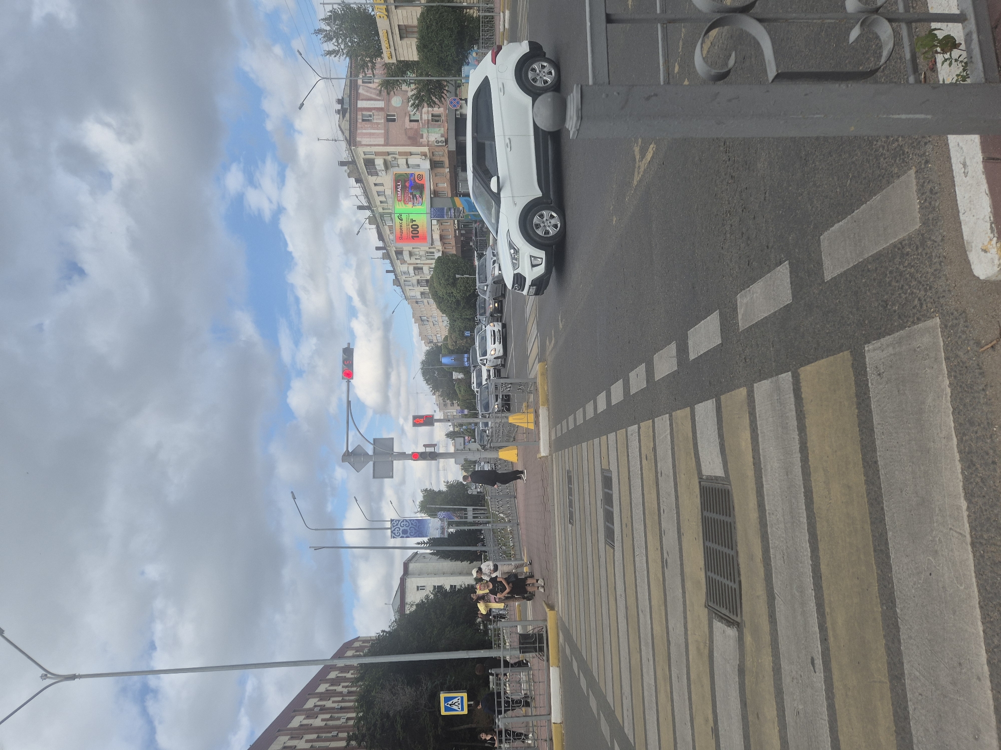 The image depicts a street scene in Kokshetau, Kazakhstan, with a pedestrian crossing in the foreground. Several people are visible on the sidewalk, waiting to cross or walking. Traffic is moving on the road, including cars and a pedestrian signal. Buildings line the street, and a large billboard is visible in the background. The sky is cloudy, suggesting overcast weather. The pedestrian crossing is marked with yellow and white stripes.