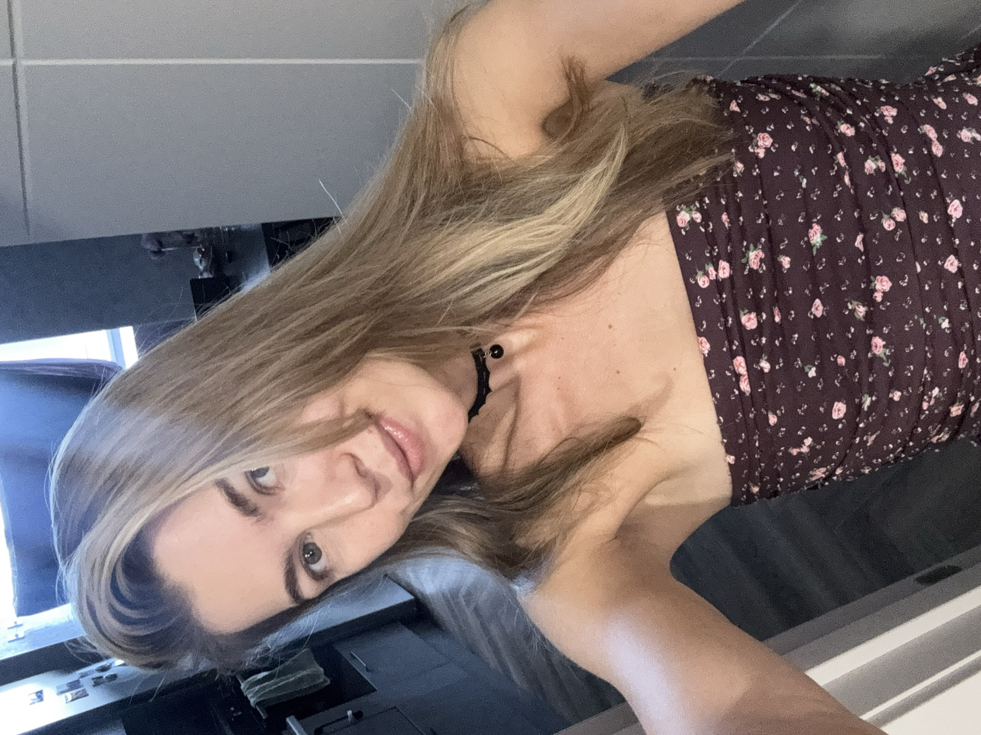 A young woman with blonde hair is taking a selfie in what appears to be a kitchen. She is wearing a strapless dress with a floral pattern and a black choker necklace. The background shows kitchen cabinets, appliances, and a tiled floor. The lighting is natural, coming from a window or overhead light.