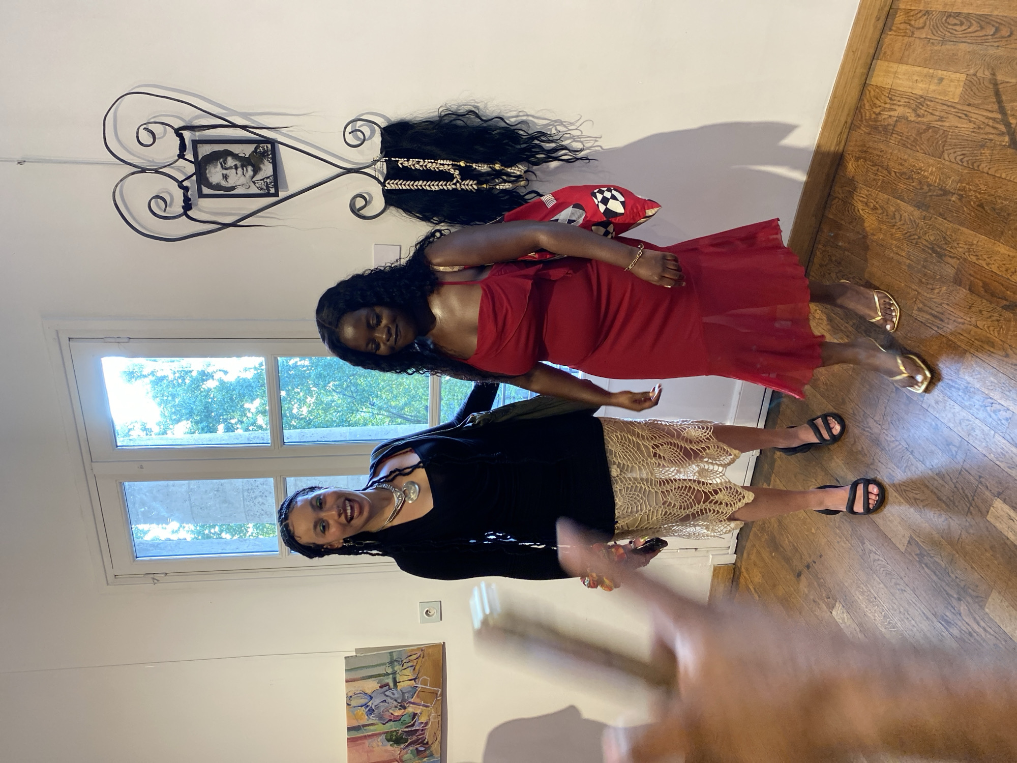 Two women are posing indoors against a wall. One woman is wearing a red dress and gold sandals, while the other is in a black top and a crocheted-style skirt with black sandals. They are standing in front of a window that looks out to green trees and a blue sky. On the wall, there is an ornate iron decoration holding a framed black and white portrait. There is also a piece of dark hair with shells attached to it and a red pillow with a checkered design. The floor is wooden. A person's hand is partially visible in the foreground, holding a phone. The location appears to be an indoor space, possibly in Paris, France.