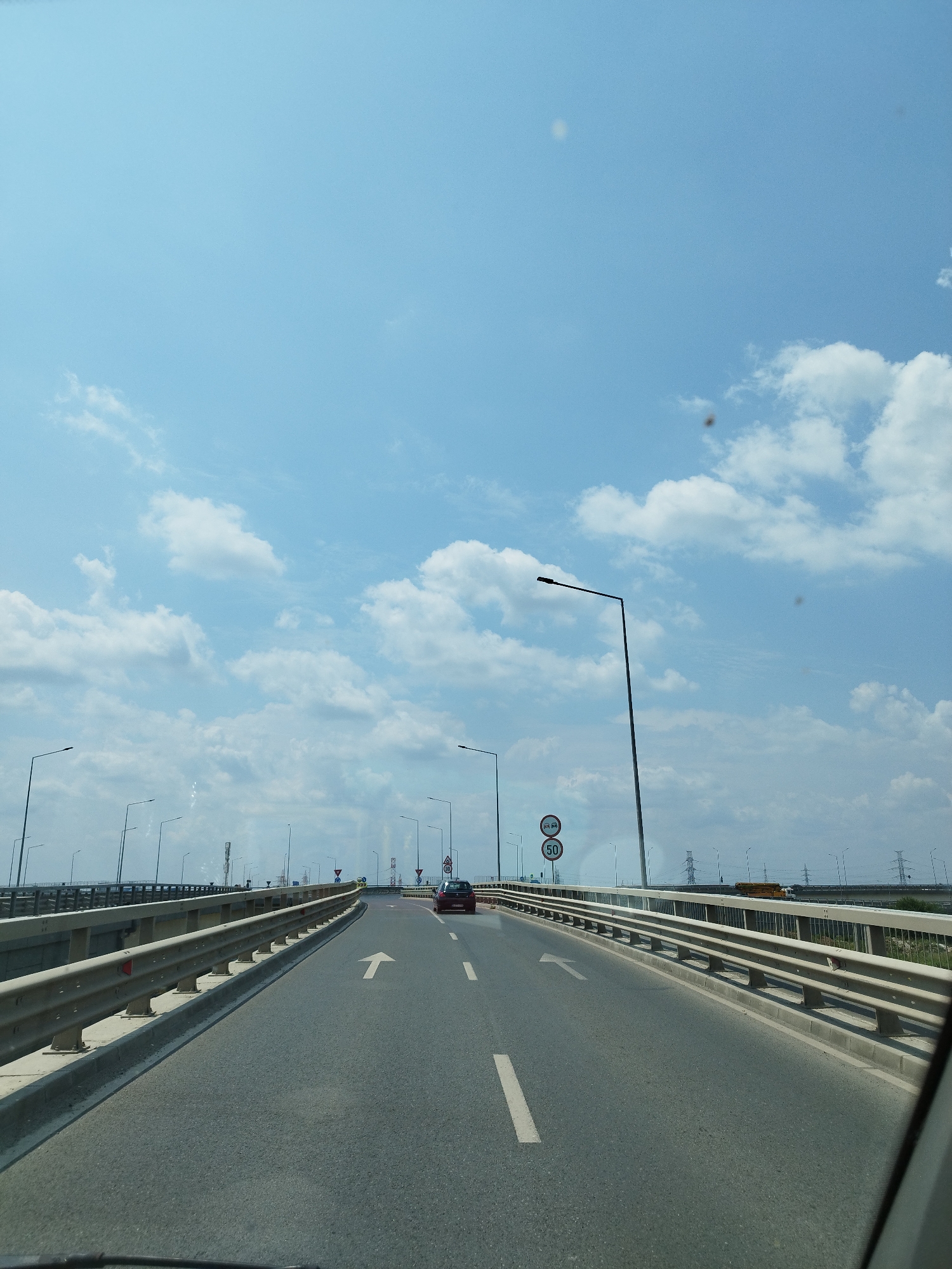 The image shows a view from a vehicle driving on a highway bridge. The sky is blue with scattered white clouds. The road is asphalt with white lane markings. There are metal guardrails on both sides of the road. Streetlights are visible along the bridge. In the distance, there are power lines and some buildings. A red car is visible in the right lane ahead. A speed limit sign indicating 50 km/h is visible on the right side of the road. The scene is set in the city of Olteni, Romania.