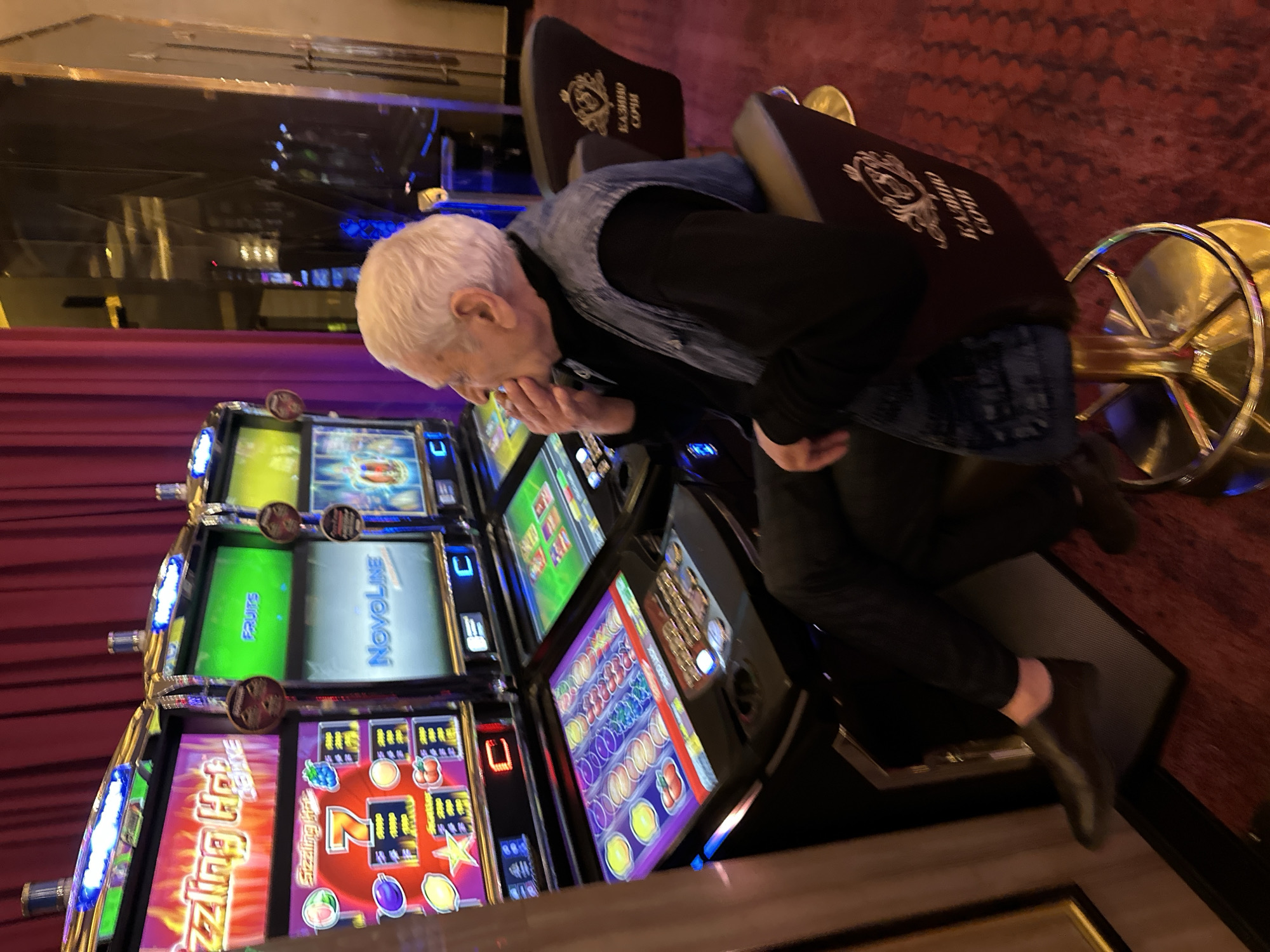 An elderly Caucasian man with white hair, wearing a black turtleneck, a dark denim vest, and black pants, sits in a casino chair. He is positioned in front of a row of slot machines, one of which displays the title "Sizzling Hot Deluxe." The man is holding what appears to be a cigarette to his mouth with his right hand and is looking to his left. The casino has a red carpeted floor and purple velvet curtains in the background. The chair he is sitting on has a logo that reads "КАЗИНО СОЧИ," indicating the location is Casino Sochi in Estosadok, Russia. The slot machines are brightly lit, with screens displaying various game graphics and text. The overall scene depicts a moment of gambling in a casino setting.