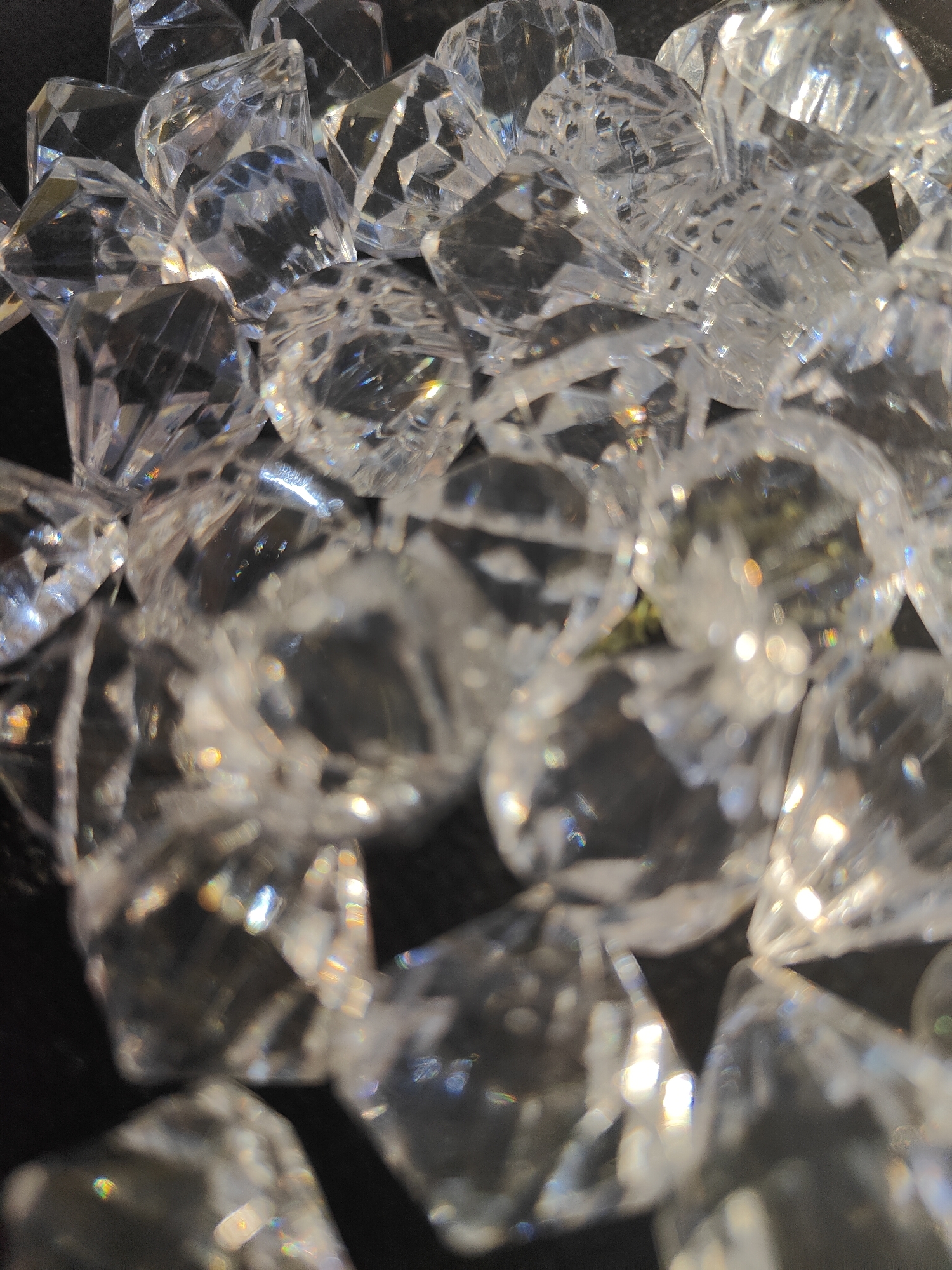 A close-up, top-down view shows a pile of clear, faceted crystal-like objects on a dark surface. The objects are irregular in shape and size, with many sharp angles and flat surfaces that reflect light. They appear to be artificial gemstones or decorative pieces, possibly made of glass or acrylic. The lighting creates bright highlights and subtle rainbow refractions within the crystals, suggesting a light source is present and illuminating them from above. The background is a solid dark color, likely black, which makes the translucent and reflective nature of the crystals stand out. The image is slightly blurred in some areas, particularly towards the bottom and edges, giving a shallow depth of field effect. There is no indication of people, specific time of day, weather, or location. The overall impression is one of sparkle and light play, focusing solely on the texture and form of the crystalline objects.