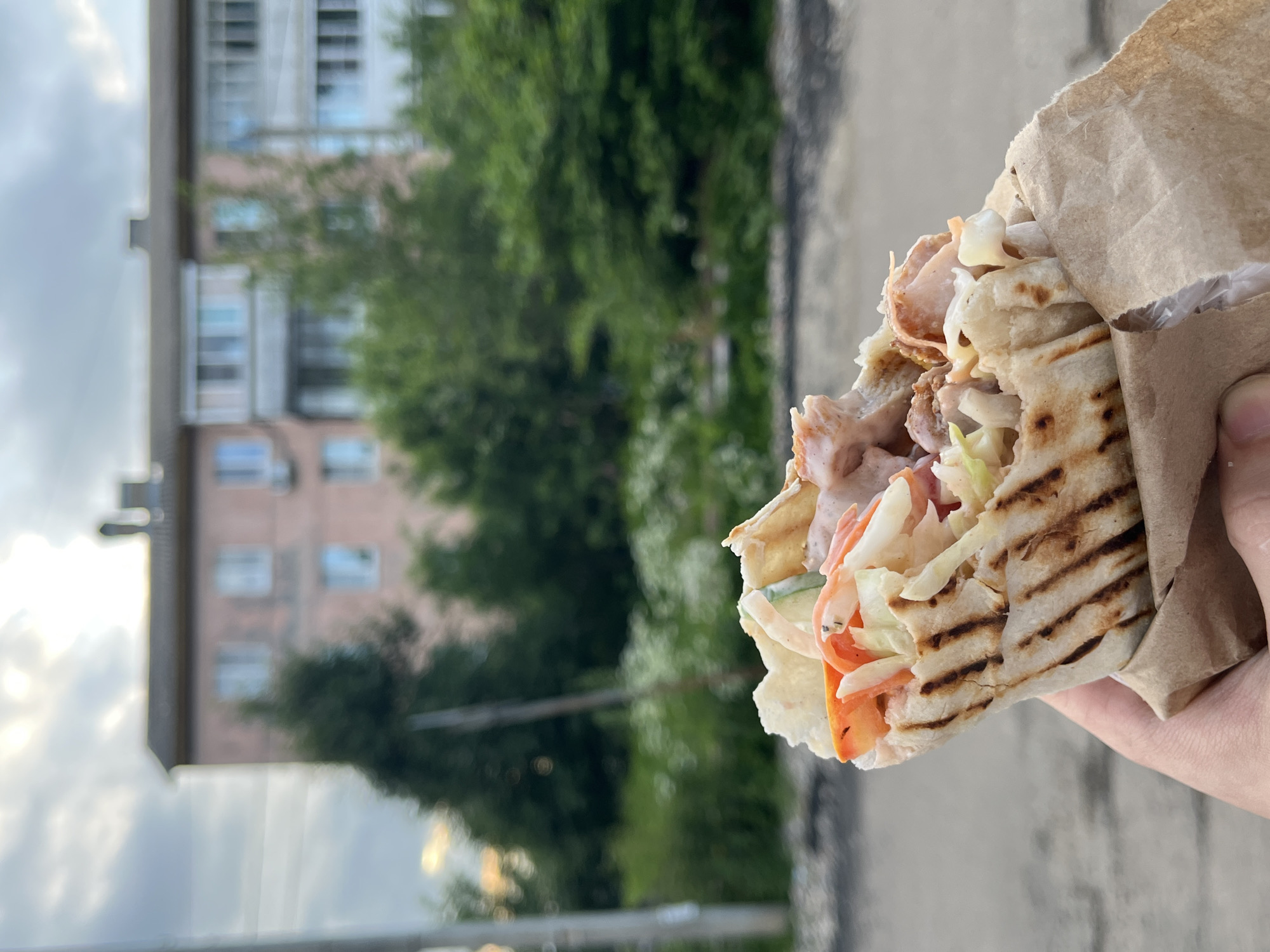 A hand holds a partially eaten shawarma wrapped in brown paper. The shawarma is filled with meat, shredded lettuce, tomatoes, and what appears to be a creamy sauce. The bread is grilled, showing distinct char marks. In the background, a multi-story apartment building with balconies and windows is visible, partially obscured by lush green trees and foliage. The sky is overcast with white clouds. The scene suggests an outdoor setting, possibly in a residential area, given the presence of the apartment building. The lighting indicates it is daytime, likely afternoon given the soft light. The overall mood appears casual and everyday. No visible text is present. The location is identified as Nikolskoye, Russia.