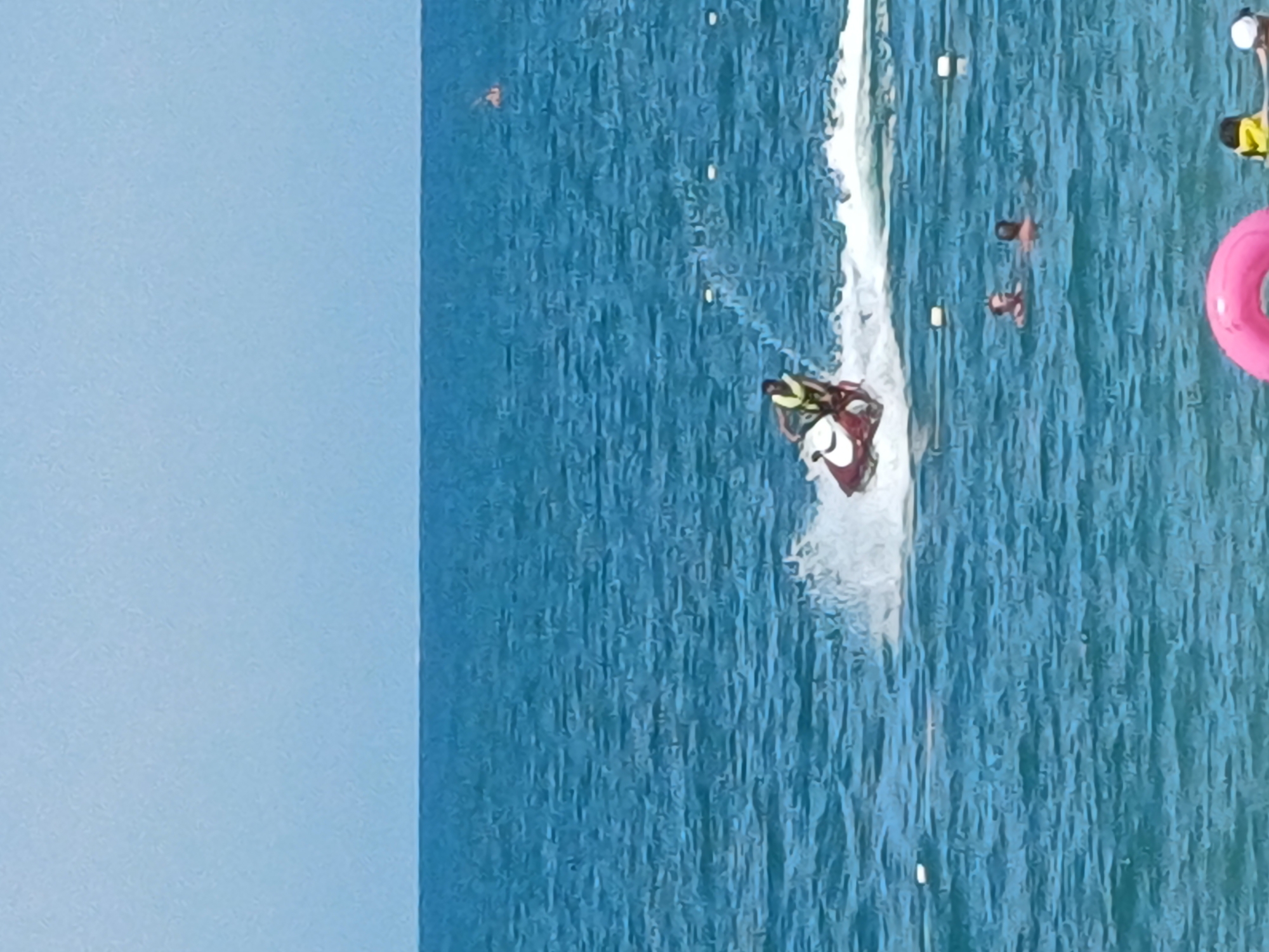 This is a horizontal, eye-level shot of a beach scene on a sunny day. The water is a deep blue and appears choppy with white caps. In the mid-ground, a person on a red and white jet ski is speeding through the water, creating a large spray. Further back, a few people are swimming and wading in the ocean. A pink inflatable ring is visible in the foreground on the right. In the far distance, a small orange buoy bobs in the water. The sky is a light, hazy blue. The location is a beach in the city of Sochi, Russia.