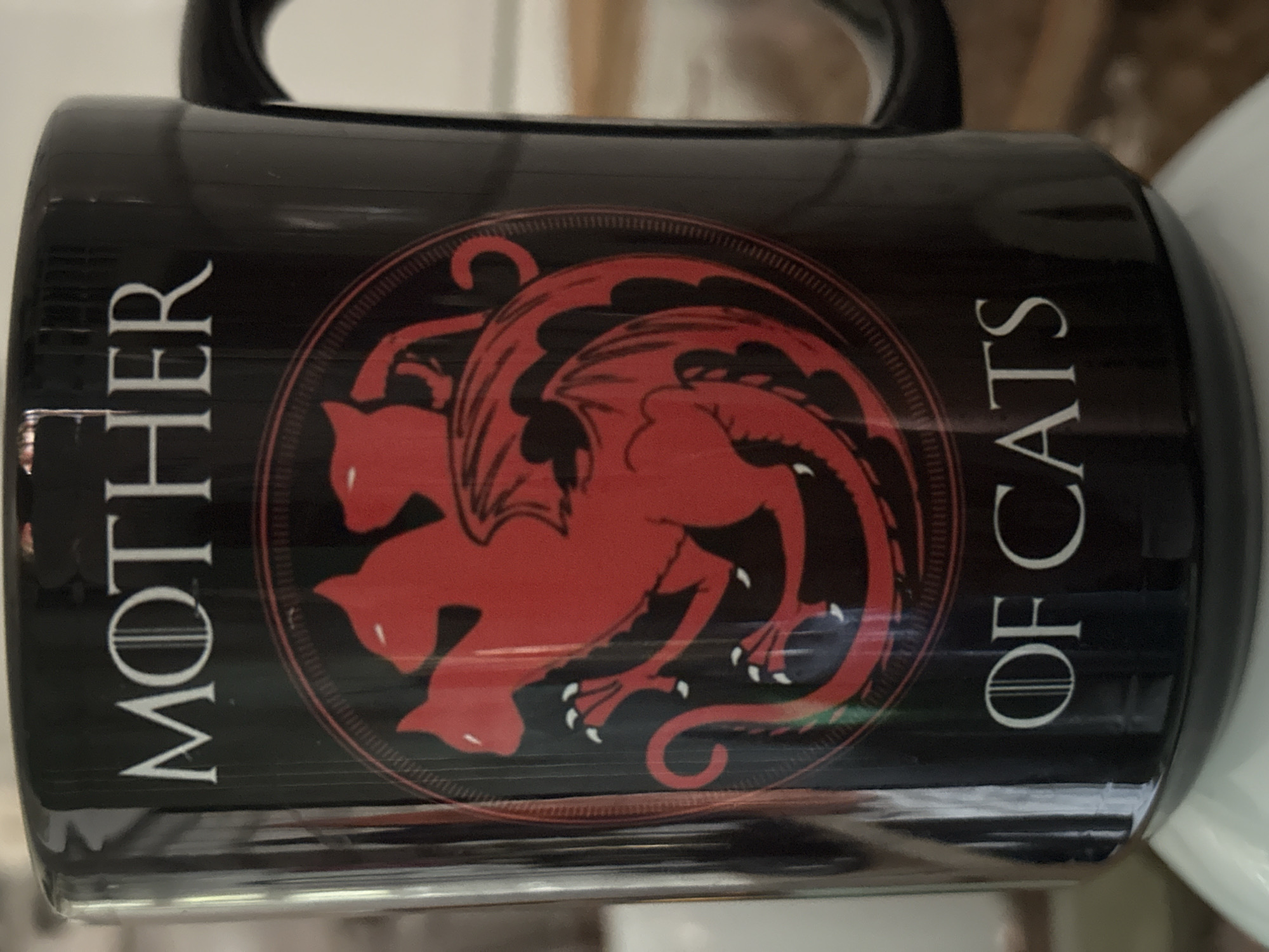 The image shows a black mug with a red design featuring three stylized cats arranged in a circle, resembling a dragon. The words "MOTHER" and "OF CATS" are printed in white, vertically aligned on opposite sides of the design. No location information is available in the image.