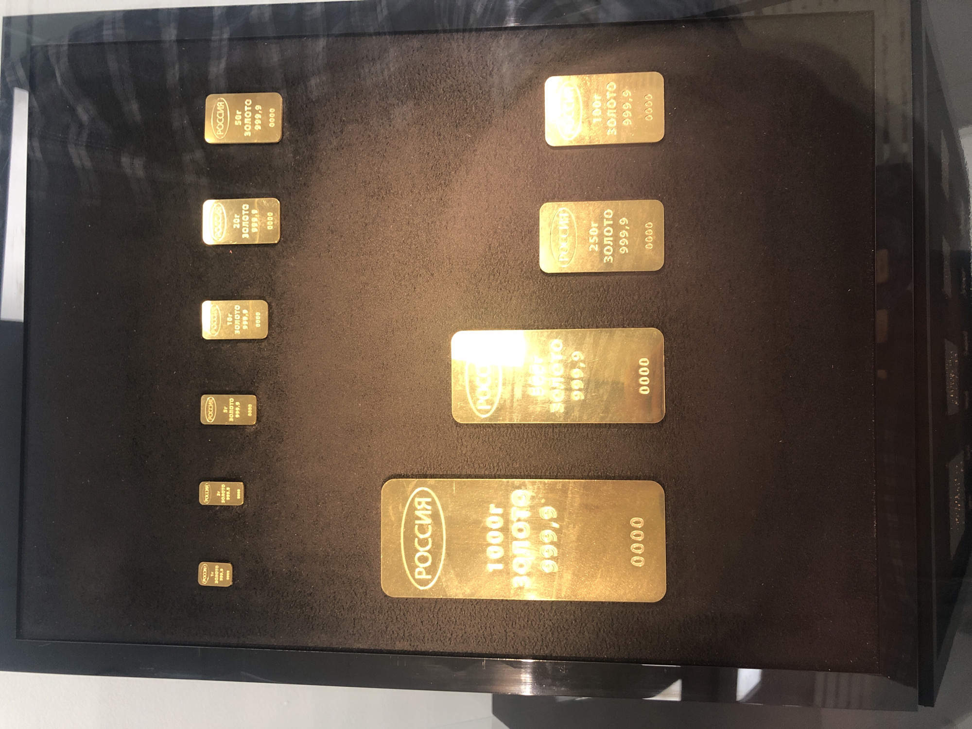 A collection of gold bars of varying sizes is displayed in a black box frame. The bars are arranged on a dark background, increasing in size from top to bottom and from left to right. Each bar is engraved with the word "РОССИЯ" (Russia), the weight in grams (e.g., 1000г, 250г, 100г, 50г), the word "ЗОЛОТО" (Gold), and the purity "999,9." The word "0000" is also engraved on each bar. The bars are neatly arranged in rows and columns, with the smallest bars in the upper left corner and the largest bar in the lower part of the frame. The location and specific details about the city and environment are unknown.