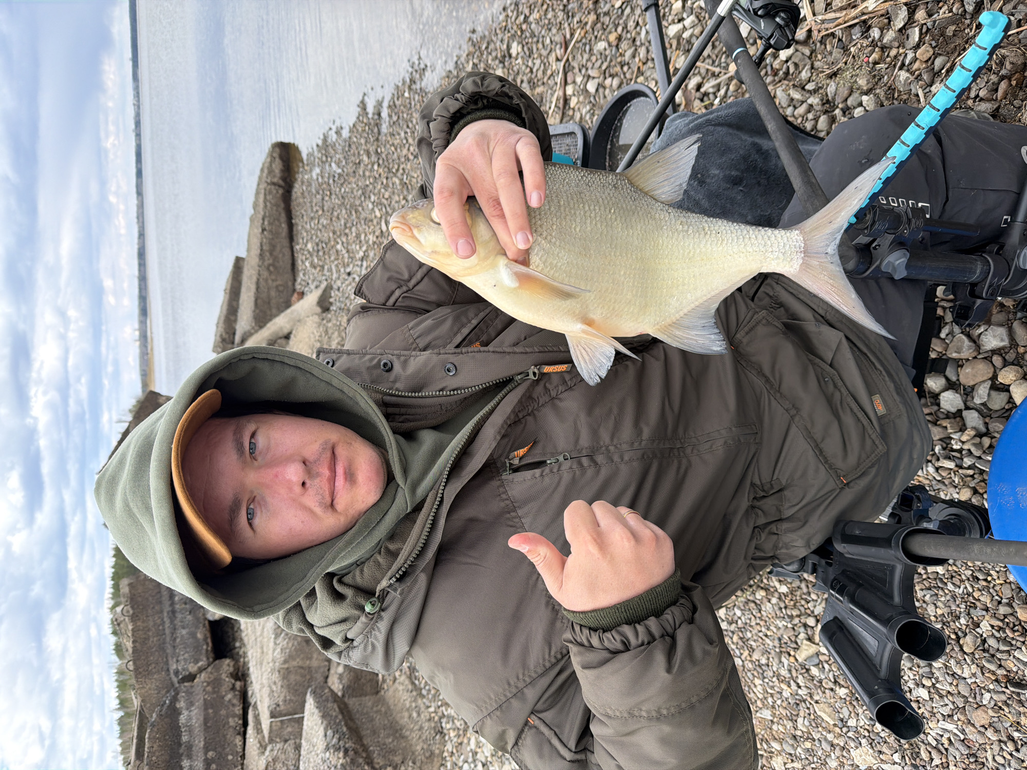 The image, rotated 90 degrees clockwise, shows a man outdoors, presumably fishing, by a large body of water. He is positioned in the foreground, looking directly at the viewer with a neutral expression, and gives a thumbs-up gesture with his left hand. The man wears a dark olive-green hooded winter jacket over a lighter green hooded layer, with a tan cap visible beneath the hood.
He is holding a light-colored, broad-bodied fish, likely a Bream, with his right hand, showcasing his catch. The fish has noticeable scales and well-defined fins. The setting is a rocky, pebbly shoreline, which extends into the midground. To the man's left, large, weathered concrete blocks are visible, possibly serving as part of a breakwater or an old structure along the water's edge. Beyond these structures and stretching across the background is a calm, expansive body of water, under an overcast sky with visible clouds. This environment is consistent with the location context of Chaikovsky, Russia, which borders the Kama River and Votkinsk Reservoir.
Around the man, various fishing gear is present. A black fishing bag or seat box, displaying the text "URSUS," is next to him. A fishing rod with a distinctive bright blue handle grip is also visible, along with other rod components and a blue bucket or container in the lower right. The overall scene suggests a successful fishing trip on a cloudy day by a river or reservoir.