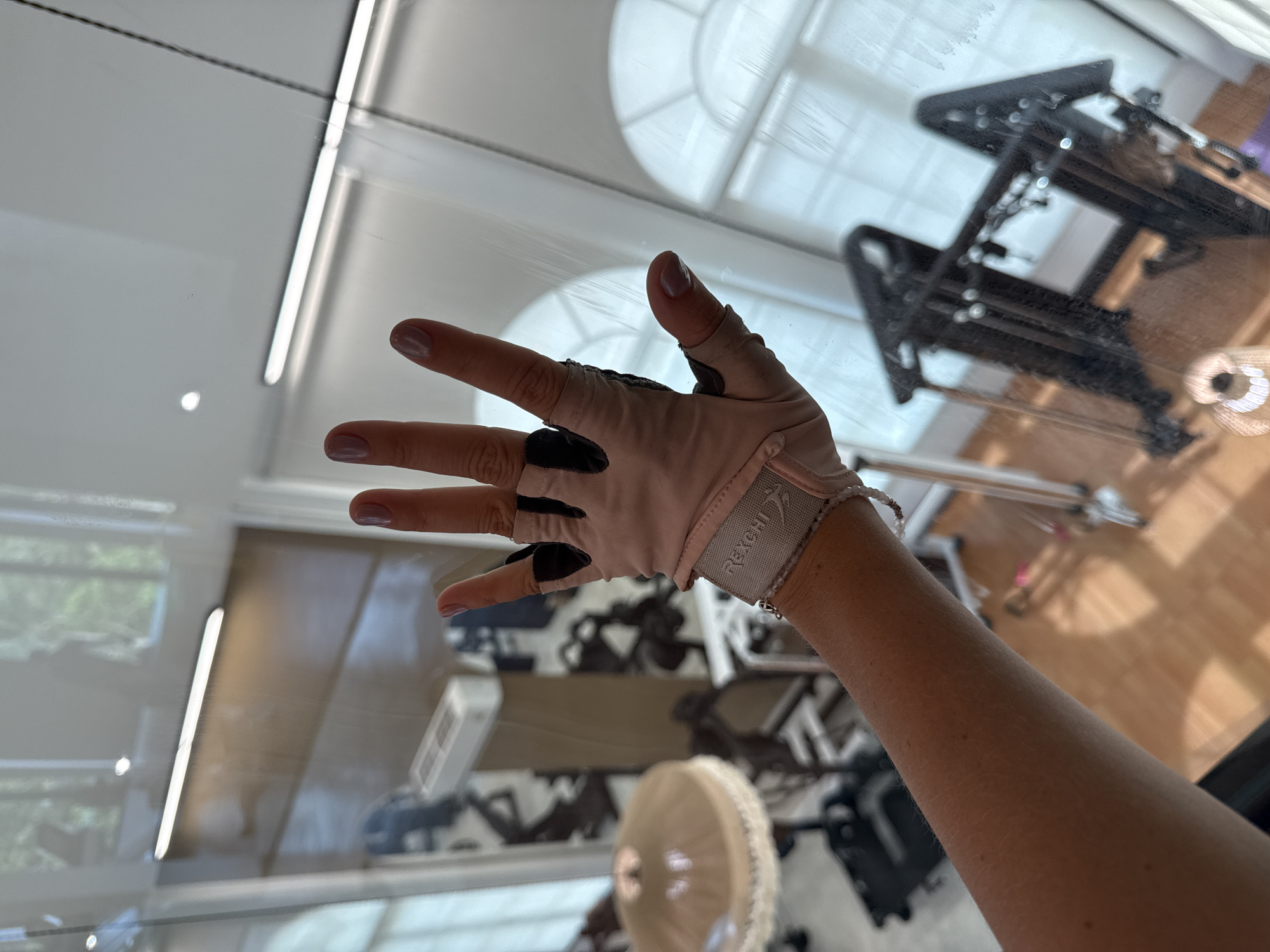 A forensic, matter-of-fact description of the scene in ตำบลเกาะพะงัน, Thailand, shows a human left forearm and hand pressed against a clear glass partition. The hand wears a light pink or beige fingerless fitness glove featuring black accents on the knuckles and a wrist strap with "REXCHI" embossed in white. A multi-strand light-colored beaded bracelet adorns the wrist. The visible fingernails are painted with a pale pink polish. Through the glass, an indoor fitness studio is visible, equipped with several dark-colored exercise machines resembling Pilates reformers. A light-colored, pleated lampshade hangs on the left. A white, rectangular air conditioning unit is mounted high on a background wall. Multiple arched windows with white frames are visible in the far background, illuminating the room with natural light and showing blurred greenery outside. The floor of the studio is composed of light-colored wooden planks. Reflections on the glass partition include a long, horizontal fluorescent light fixture and a circular recessed light, indicating overhead illumination in the area from which the photograph was taken. The glass surface exhibits subtle smudges and streaks from contact.