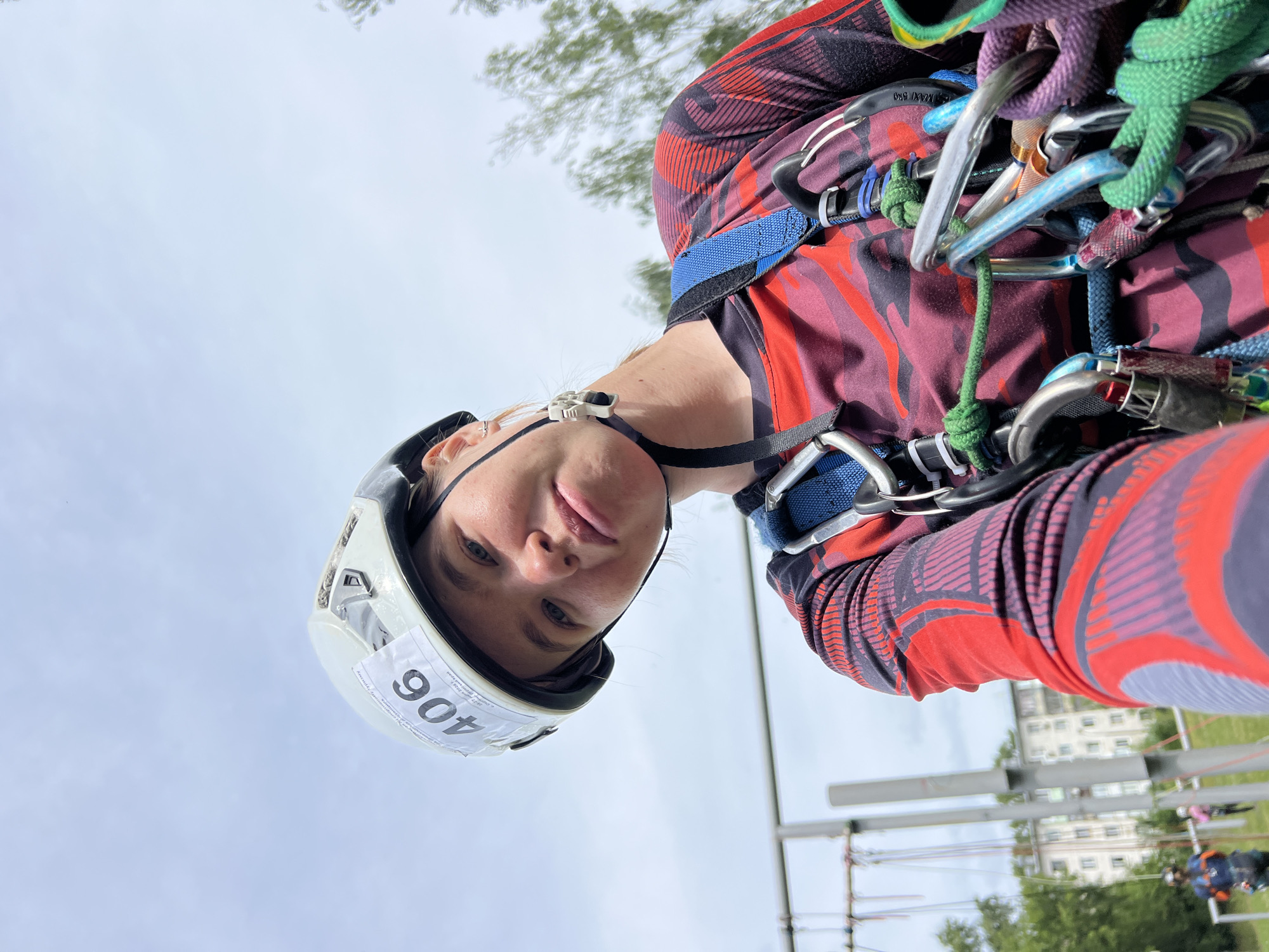 This is a vertical, close-up, selfie-style photograph taken outdoors, with the camera positioned low and looking upwards. The main subject is a young woman wearing a white helmet with the number "406" visible on a sticker attached to the front. She has blonde hair and appears to be wearing a red and purple long-sleeved shirt with a geometric pattern. Around her shoulders and chest, she is equipped with a climbing harness and various carabiners, ropes, and other climbing gear, suggesting an activity related to climbing or an obstacle course. Her expression is neutral, with her eyes looking directly at the camera.
The background is a bright, overcast sky with some trees visible at the top of the frame. In the distance, there are buildings that appear to be apartments or dormitories, and some structures that resemble part of an outdoor course, possibly with more participants blurred in the background. The scene is likely taking place during the daytime, given the bright sky.
The overall impression is one of preparation or participation in an outdoor sporting or recreational event. The image is slightly tilted, adding to the dynamic, selfie-like feel.