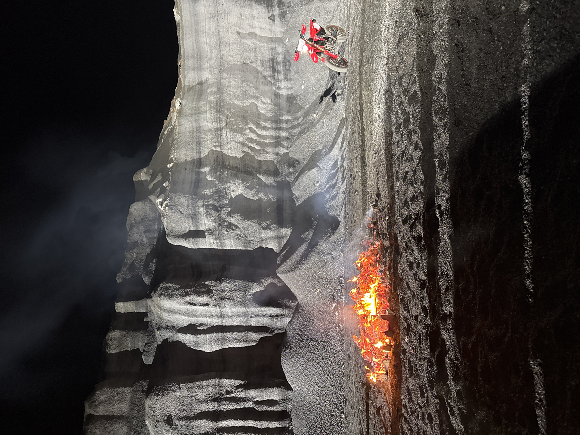 A dimly lit, vertical composition shows a motorcycle stunt taking place against a stark, layered cliff face, possibly in Teguise, Spain. The motorcycle, red and white in color, is positioned toward the upper portion of the cliff, with its rider visible but partially obscured. To the right of the motorcycle, a fire burns intensely near the base of the cliff. The cliff's surface exhibits varying textures and shadows, with distinct horizontal strata, suggesting sedimentary rock. The background is a dark, indistinct space. Smoke or mist is visible towards the left side of the cliff face.